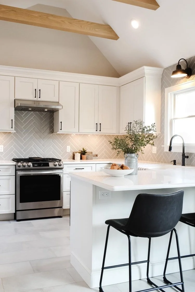 Modern white kitchen with island, black chairs, stainless steel oven, white cabinets, gray herringbone backsplash, potted plant on the counter, and window with natural light.