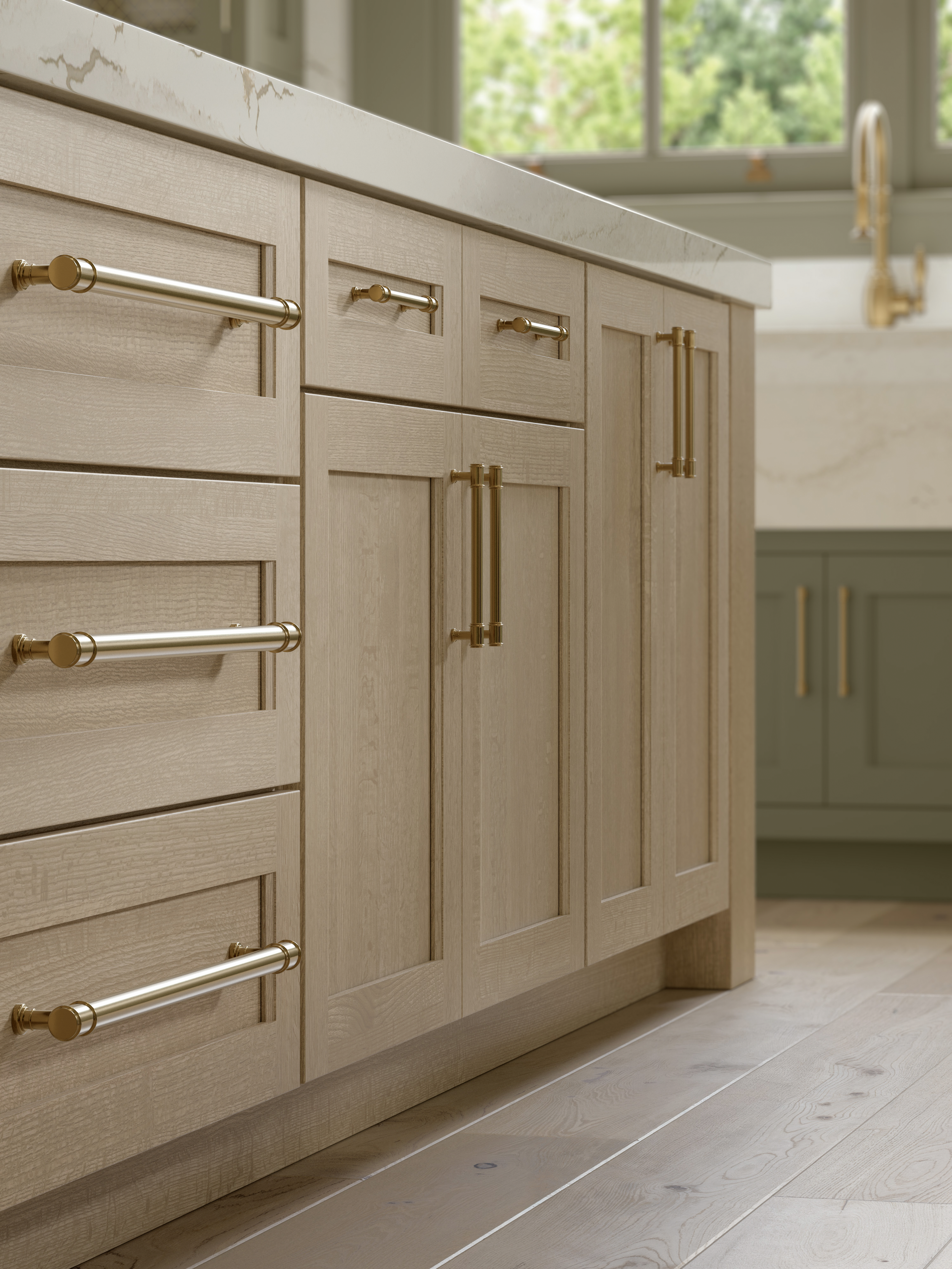 Light-colored wooden kitchen cabinets with gold hardware and a white marble countertop, with a window showing green foliage outside.