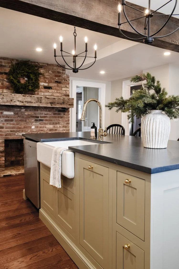 Modern kitchen island with beige cabinetry, black countertop, and gold hardware, decorated with a large white vase holding greenery, set against a brick wall with a fireplace and wooden mantel, featuring two black chandeliers and warm lighting.