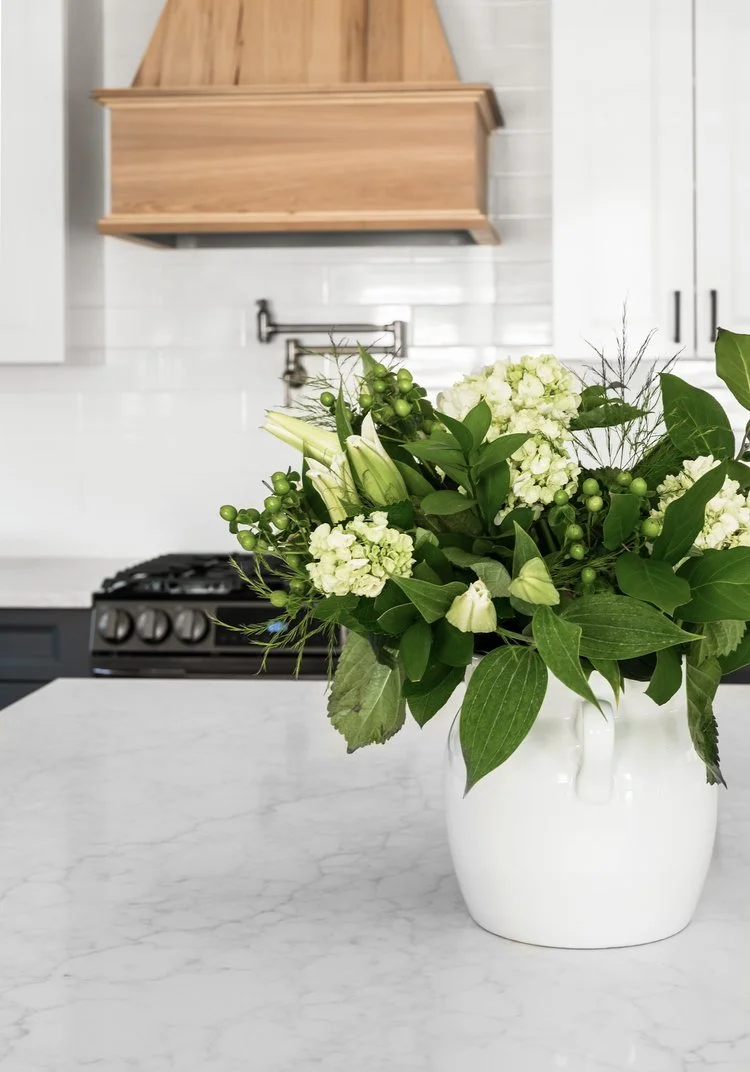 A white ceramic vase with a bouquet of white lilies, hydrangeas, and green foliage on a white marble kitchen countertop.