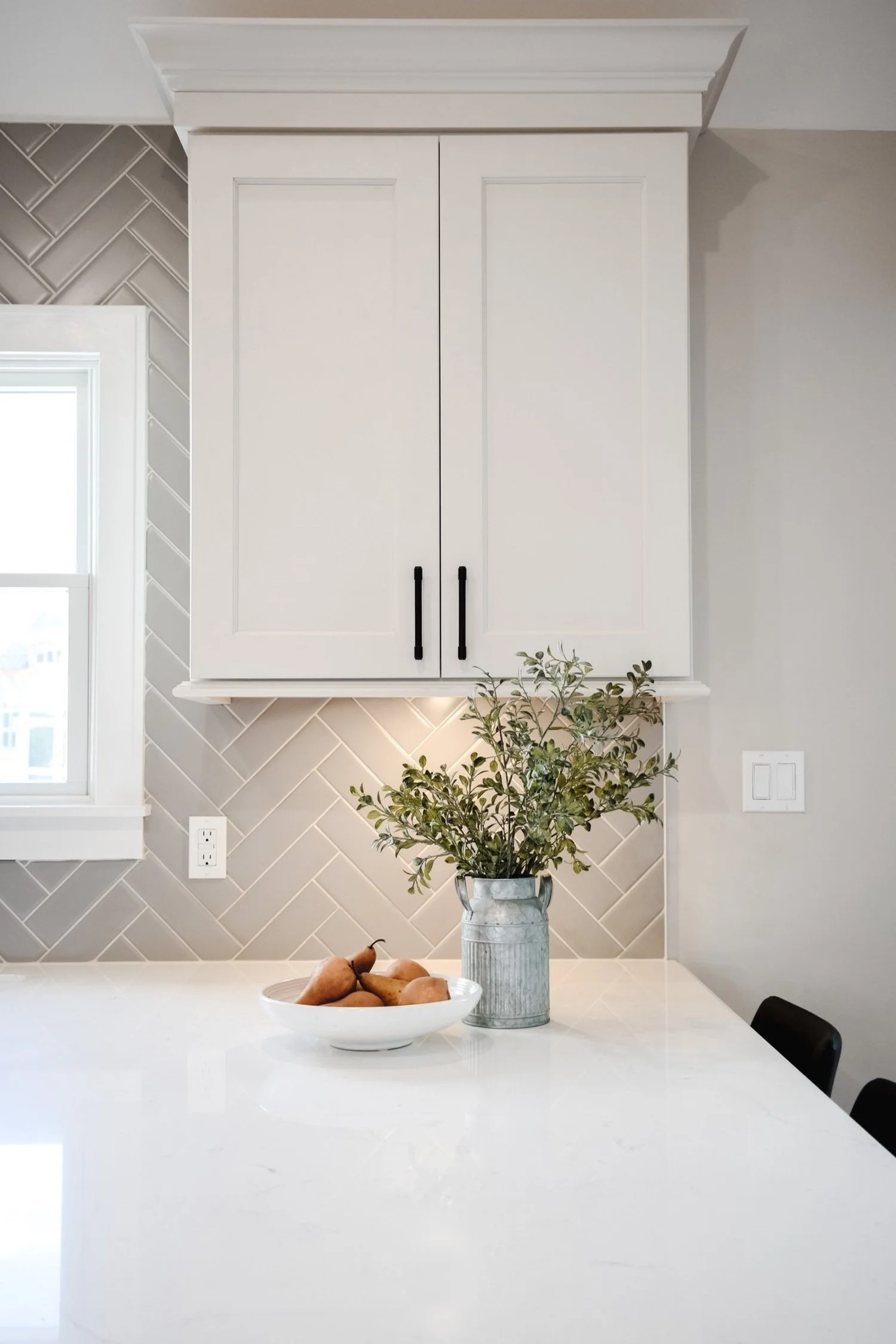 White kitchen countertop with a white bowl of pears and a vase with greenery, against a tiled backsplash and white cabinets.