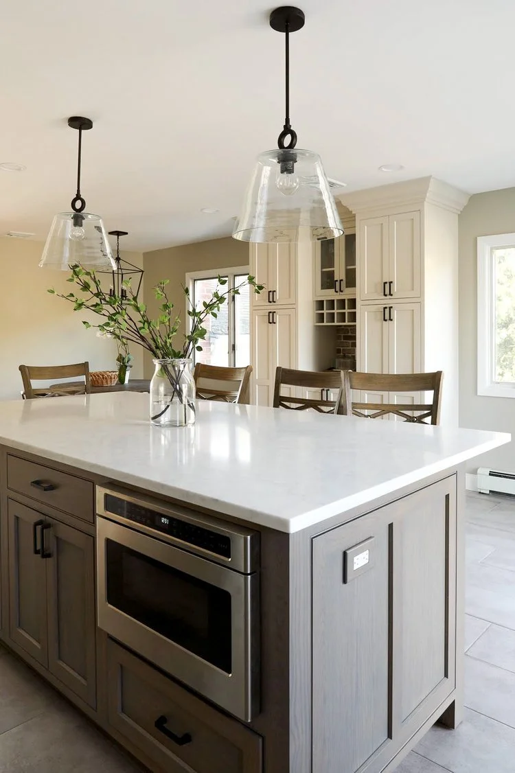 Kitchen island with a white marble countertop, a vase with green branches, hanging pendant lights, and wooden chairs in a bright, modern kitchen.