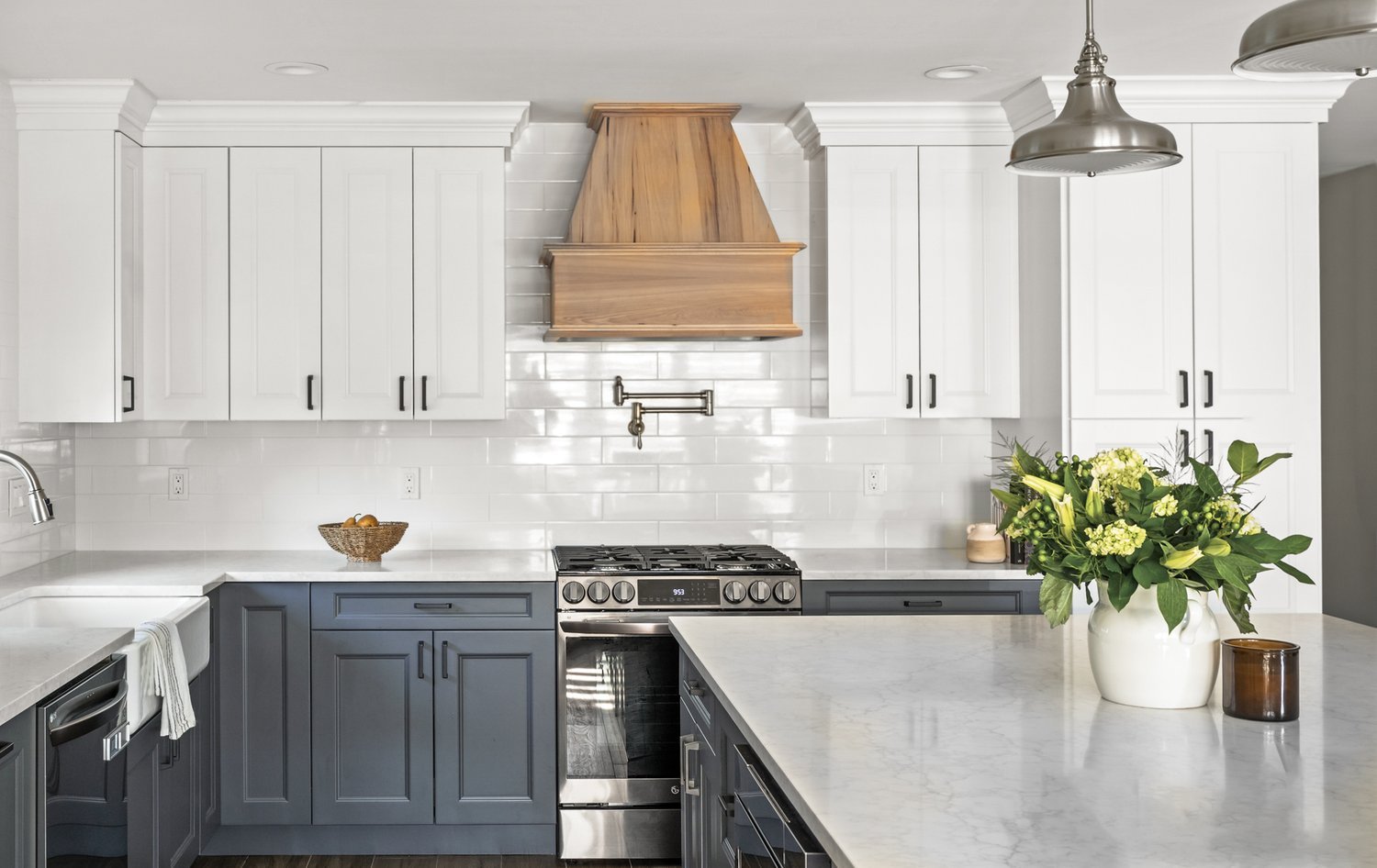 Modern kitchen with white upper cabinets, blue lower cabinets, a marble countertop island, stainless steel oven, and a wooden range hood. Decor includes a large white vase with green flowers and a dark brown container.