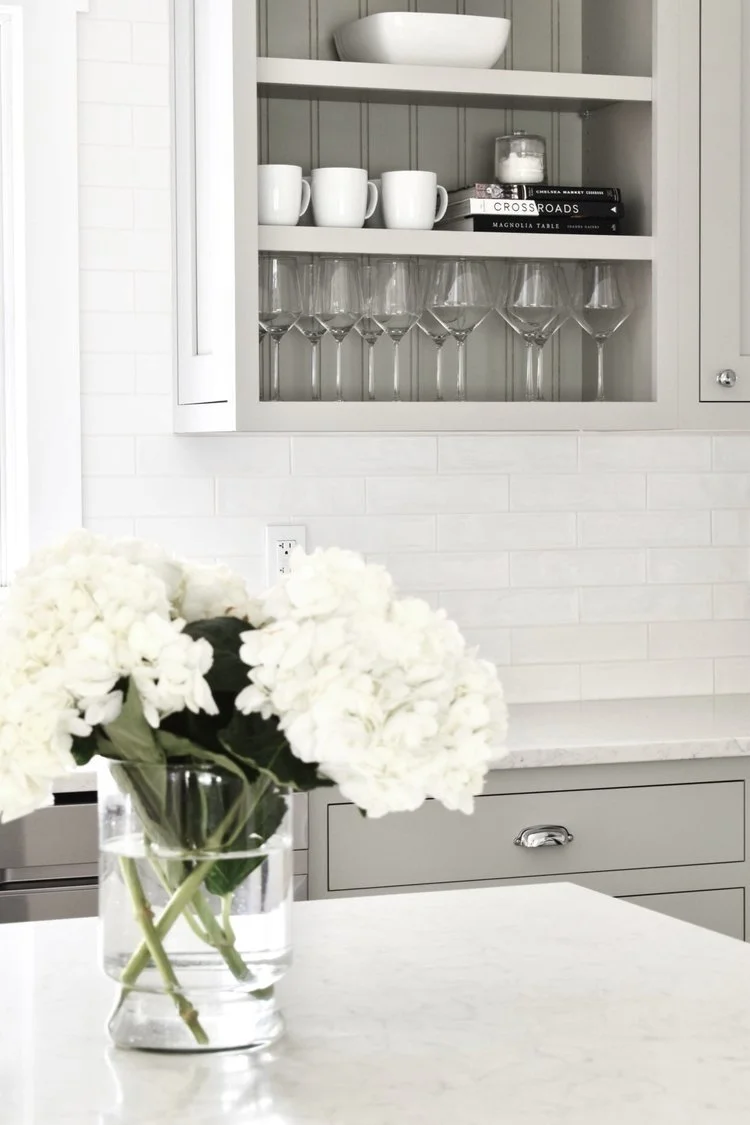 A kitchen with a gray cabinet, a white marble countertop, a vase with white hydrangeas, and a wall cabinet displaying white mugs, wine glasses, a bowl, and some books.