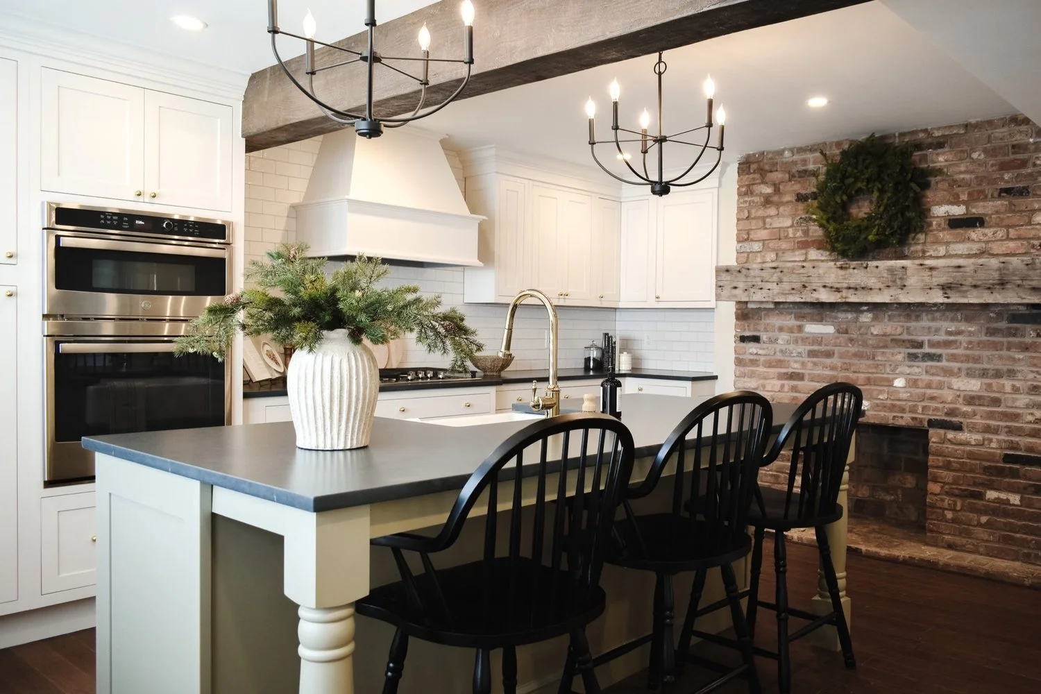 Kitchen with white cabinets, black countertop, brick wall with fireplace and a wreath, kitchen island with black chairs, large vase with greenery, chandelier lighting.