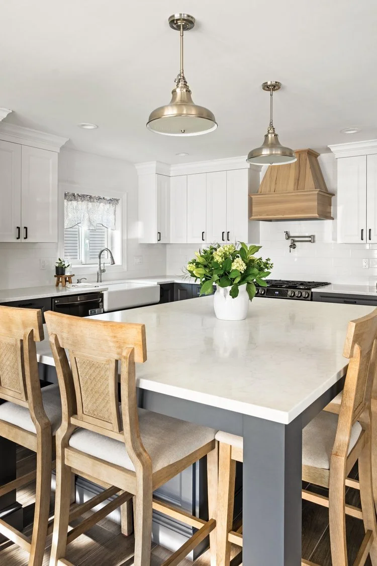 Modern kitchen with white cabinetry, a white countertop island, wooden chairs, a vase with green flowers, and pendant lights.