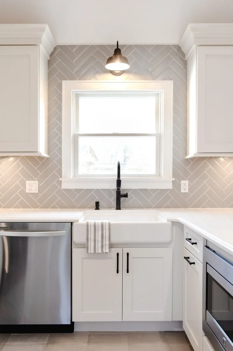 Kitchen with white cabinets, a white farmhouse sink, a black faucet, stainless steel appliances, a window above the sink, and a gray backsplash in a herringbone pattern.