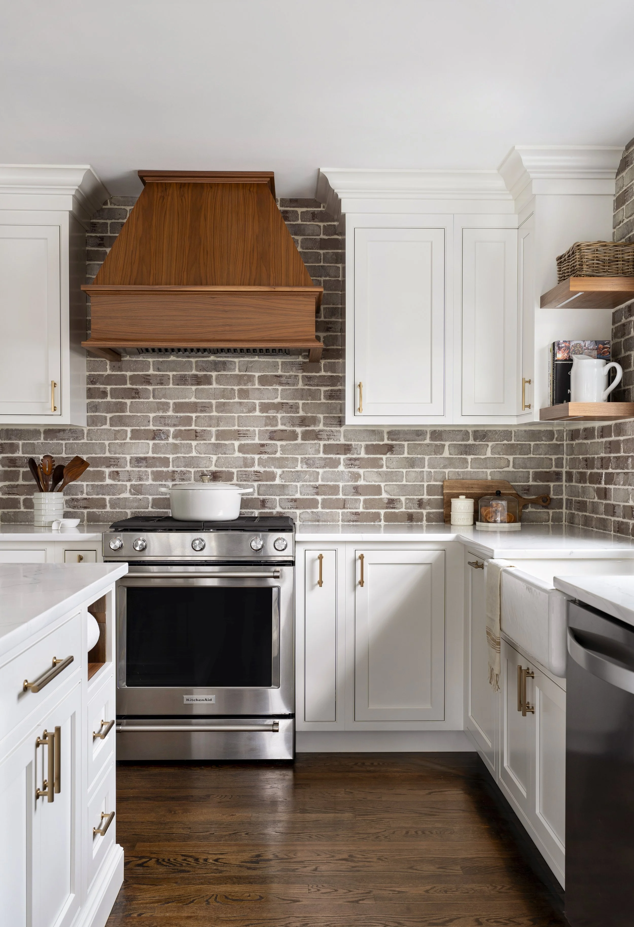 White kitchen cabinets with brass handles, stainless steel oven, brick backsplash, wooden range hood, wooden shelves, and countertop with jars and pitcher.
