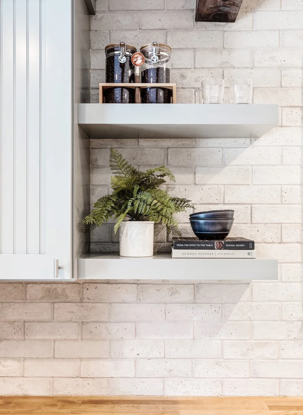 Kitchen shelf with jars of coffee beans, glasses, a potted fern, and books on a white brick wall.