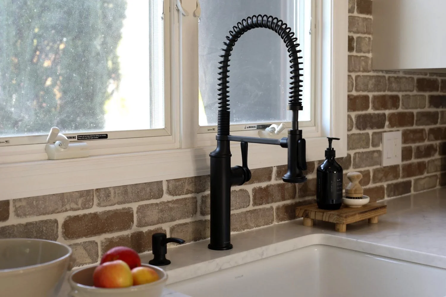 Kitchen sink area with black faucet, soap dispenser, small wooden platform with a soap dish, thyroid container, and apples in a bowl, against a brick wall and a window with mesh screen.