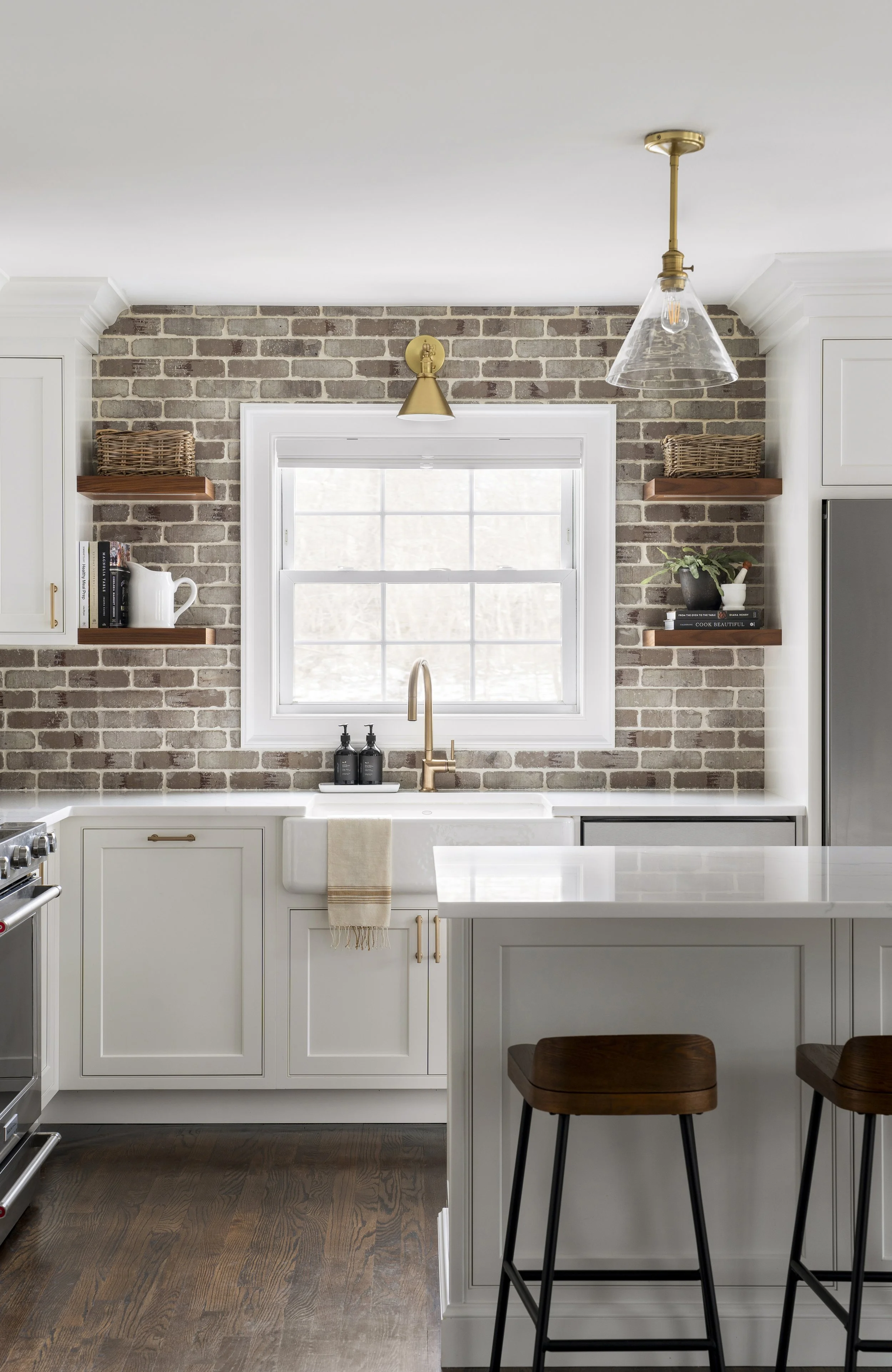 Modern kitchen with white cabinets, brick wall backdrop, window, and wooden shelves with decor and cookbooks.