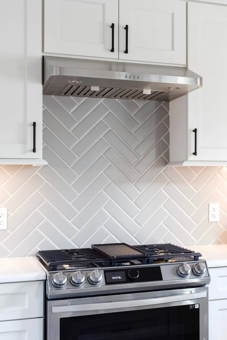 Kitchen with white cabinets, black handles, white tiled backsplash in a herringbone pattern, stainless steel range hood, and a modern stainless steel stove/oven.