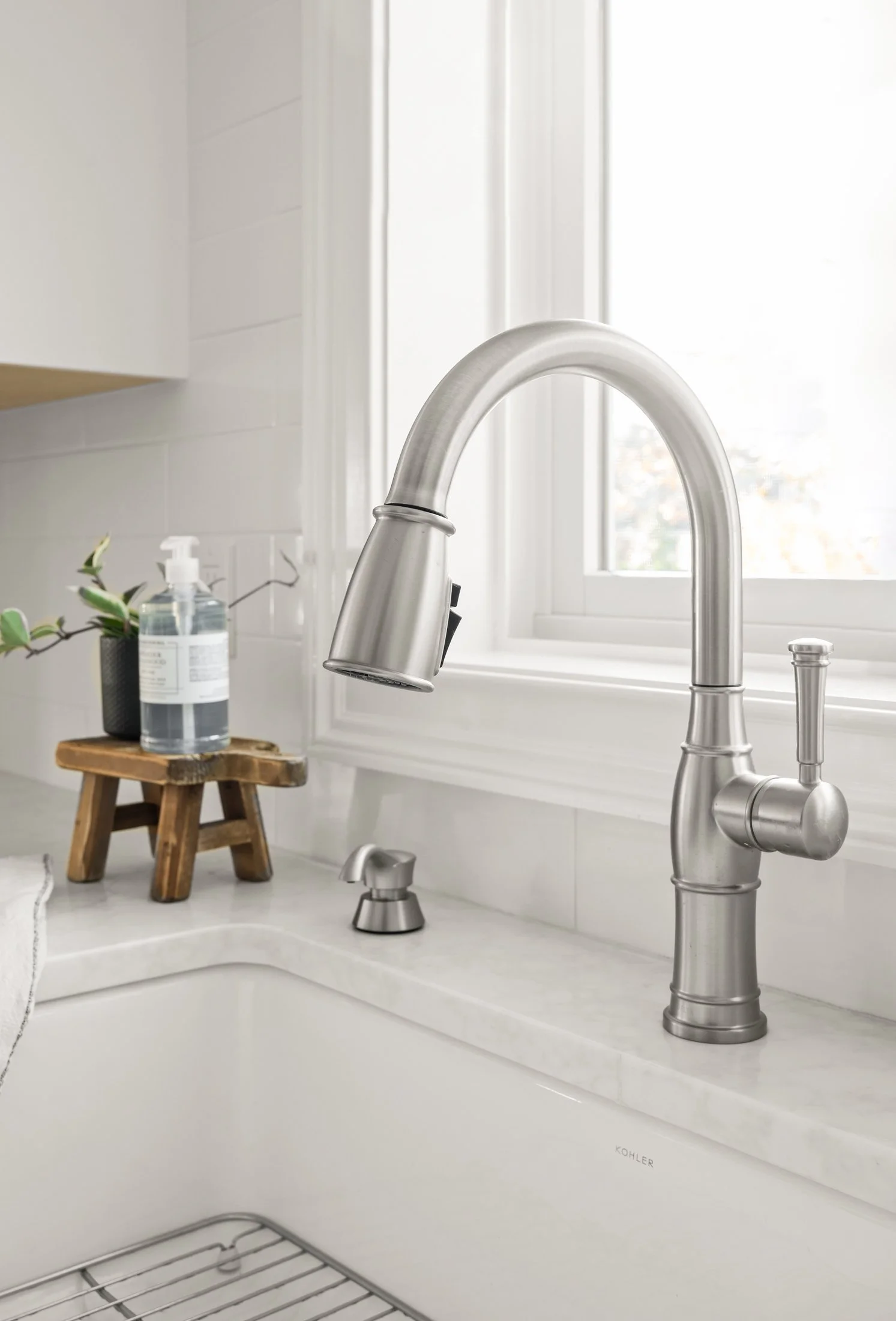 Kitchen sink with modern brushed nickel faucet, soap dispenser, potted plant, bottle of hand sanitizer, and a small wooden stool on a white countertop