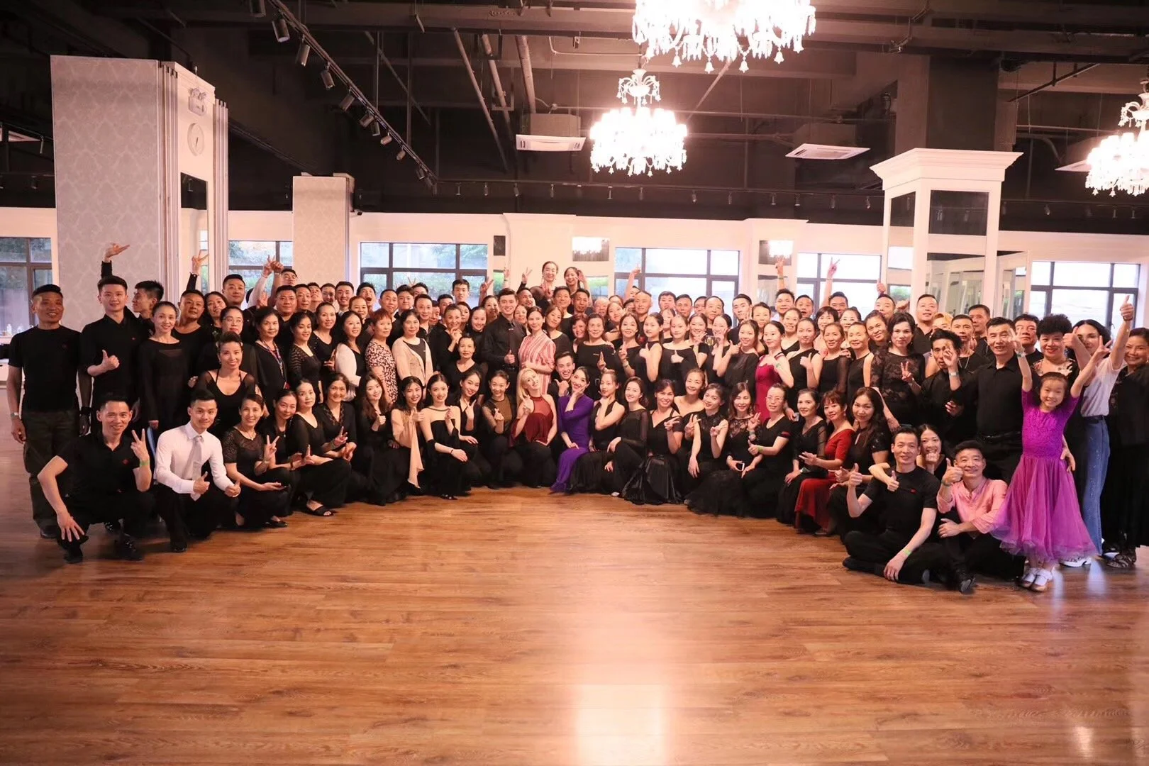 Large group of people posing for a photo in a spacious, decorated hall with wood flooring and chandeliers.