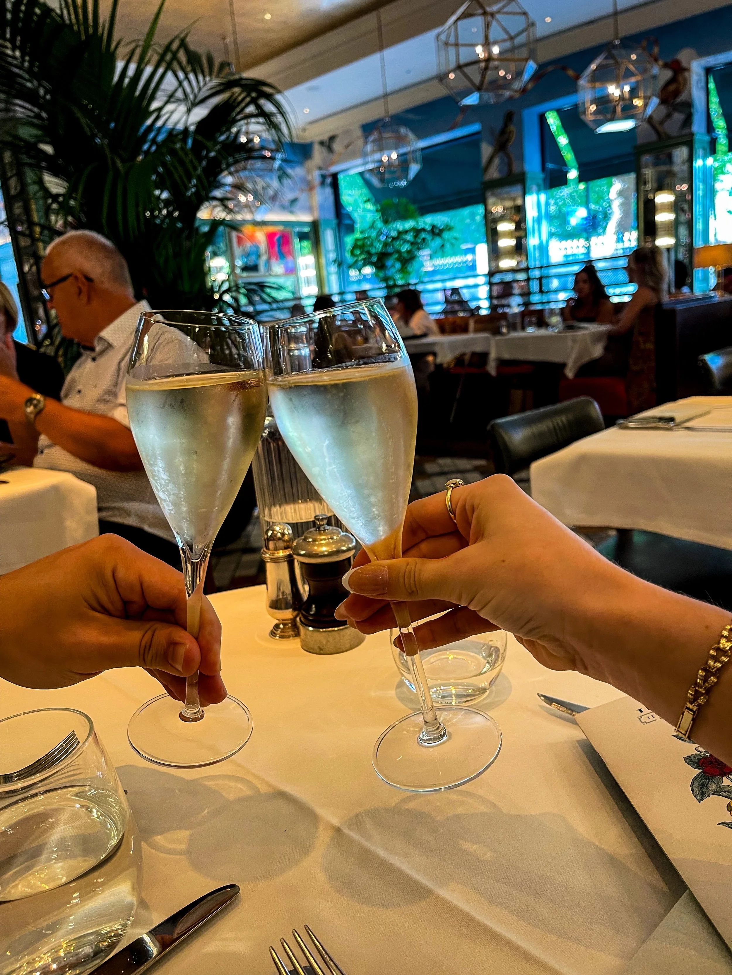 Two people clinking glasses of champagne in a restaurant with a lively, colorful interior and other diners in the background.