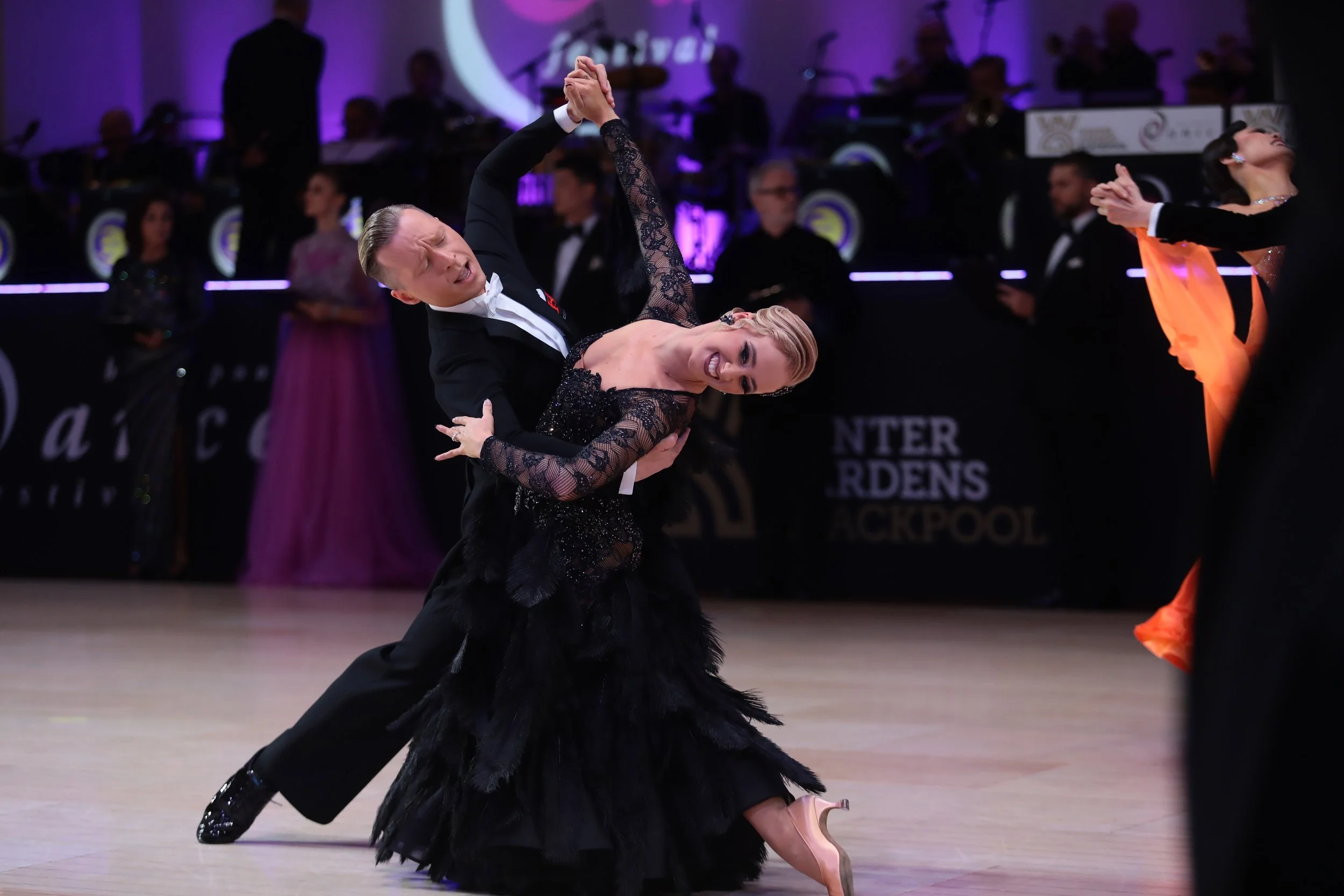 A couple performs a ballroom dance on a polished wooden floor, with the man dressed in a black tuxedo and the woman in a black, sequined gown. They are in a dramatic dip, with their hands clasped and expressive faces. In the background, musicians and a woman in a flowing purple gown are visible, along with a dark banner that reads 'Dancer's Garden Back Pool'.