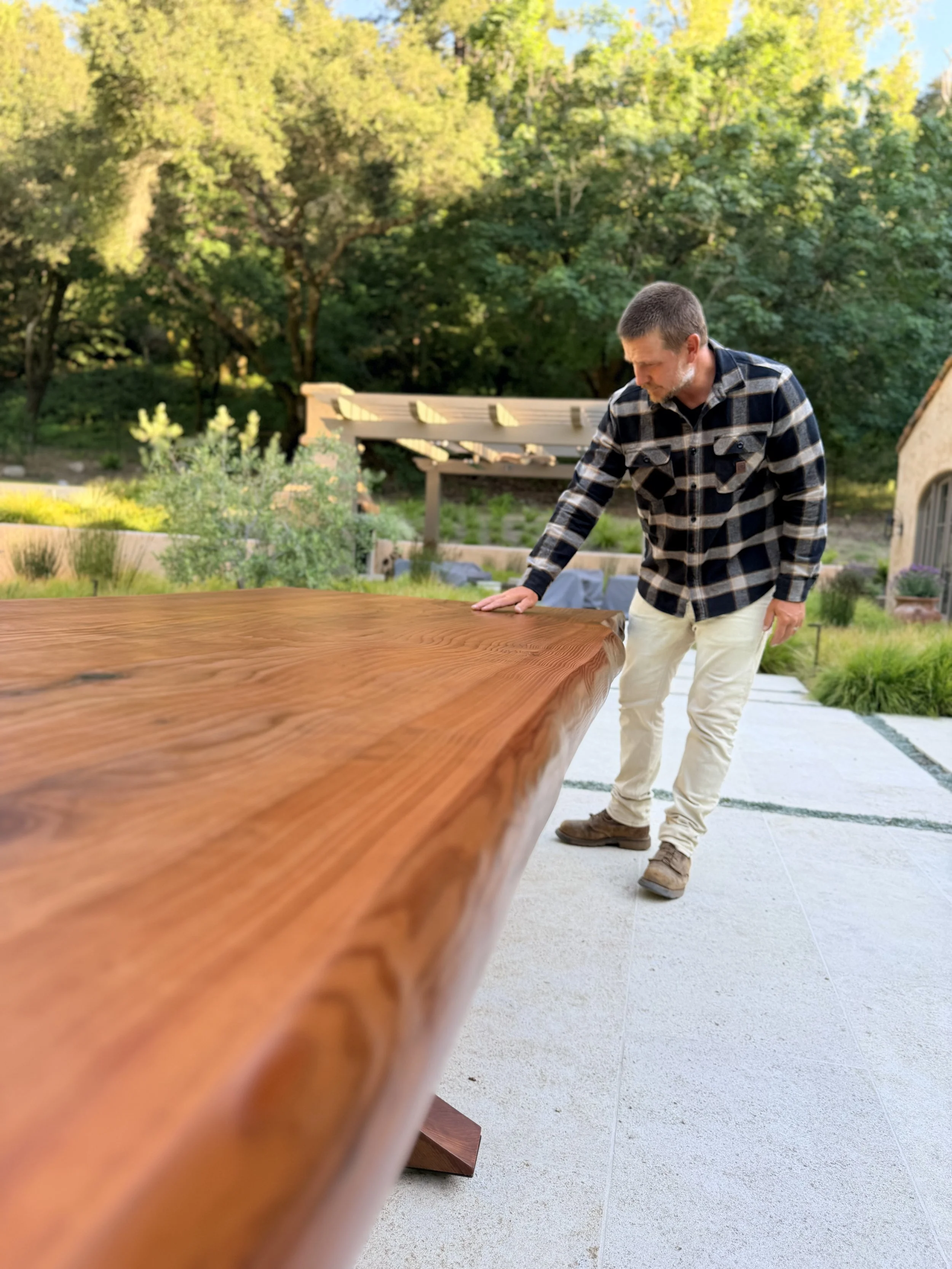 Man wearing plaid shirt and light-colored pants inspecting a long wooden table outdoors with trees and a building in the background.