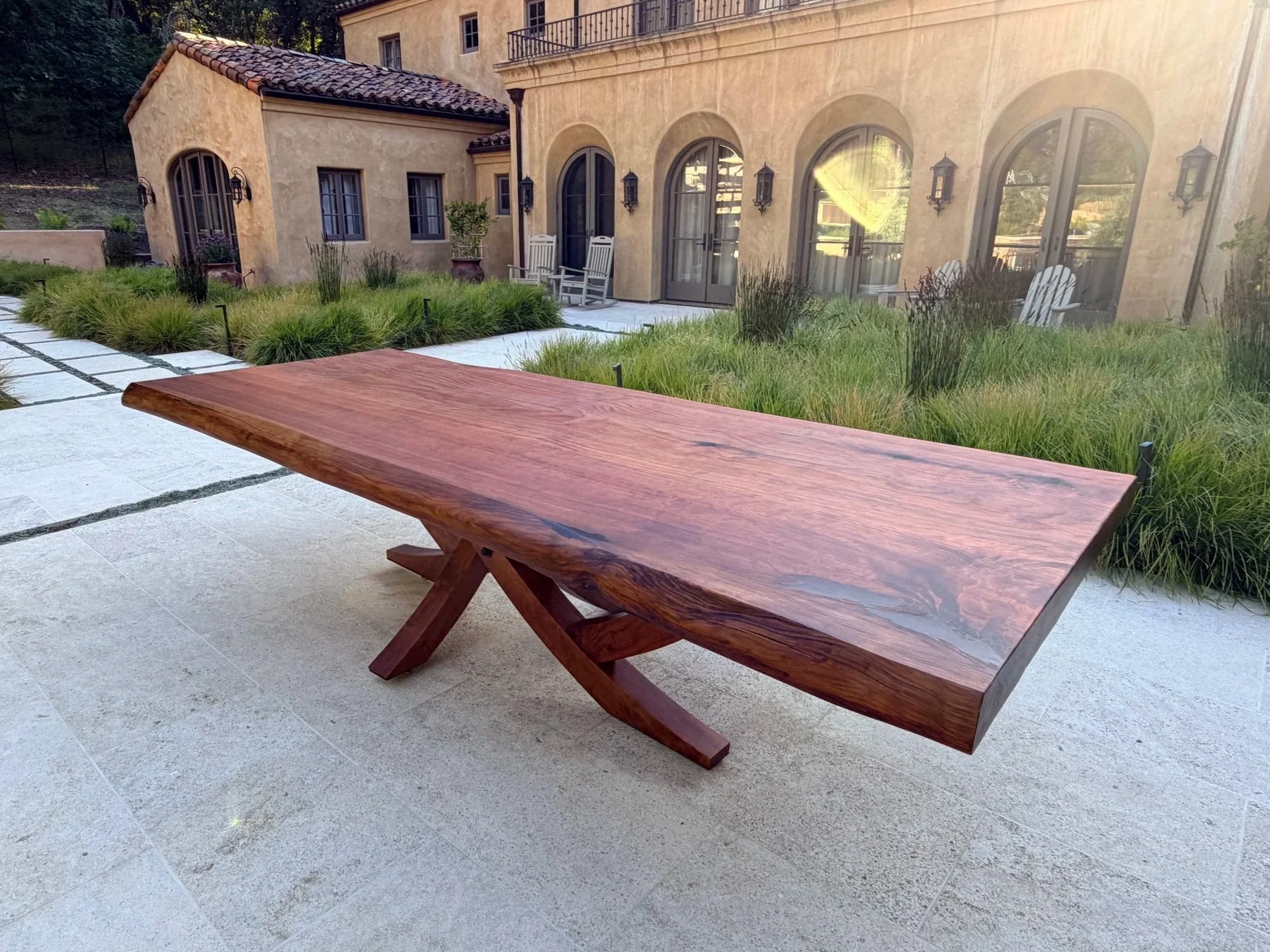 A large wooden table on a patio with a garden, house, and outdoor chairs in the background.
