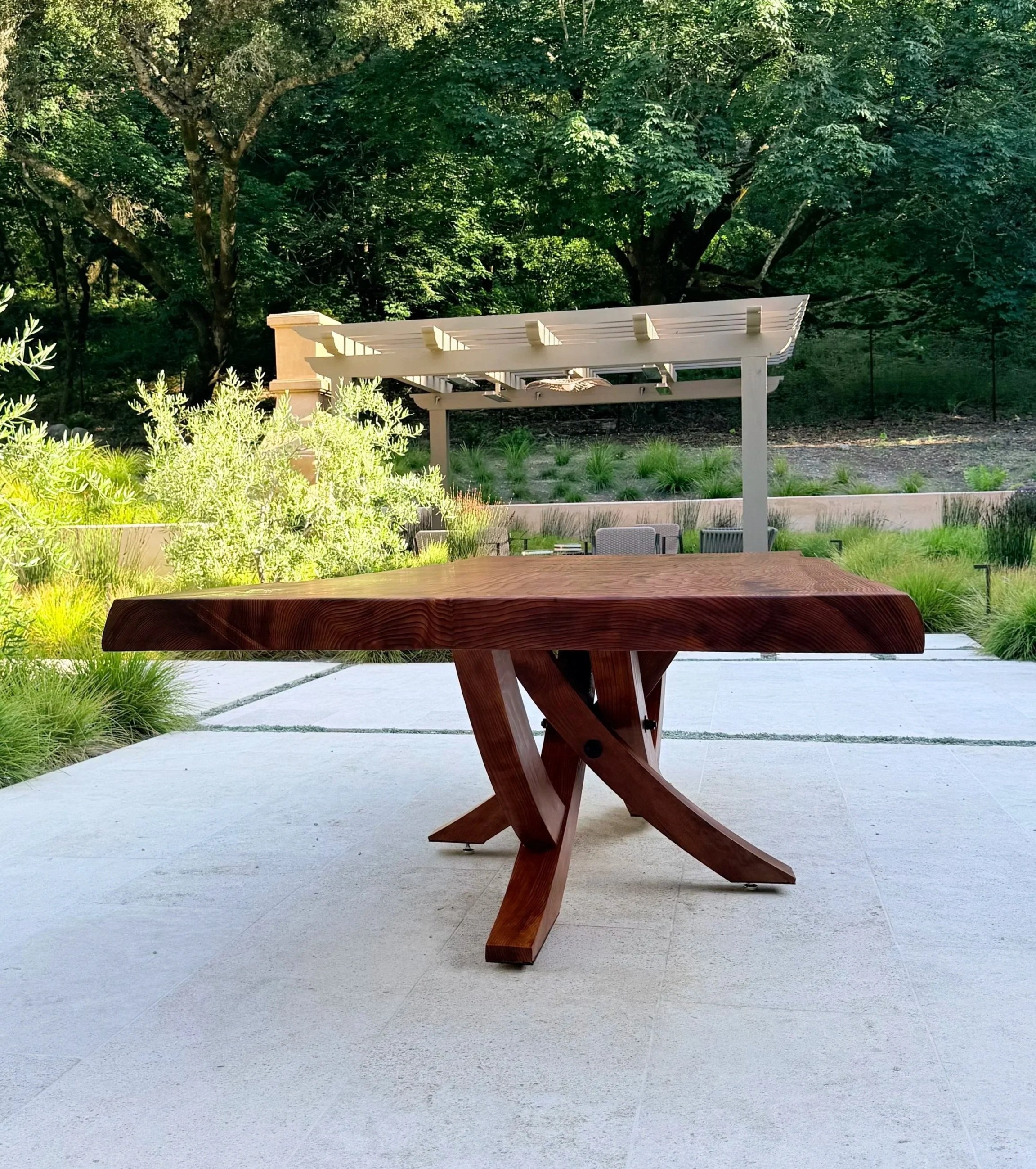 An outdoor patio with a wooden table and chairs. In the background, there is a white wooden pergola, lush green trees, and a garden with various plants.