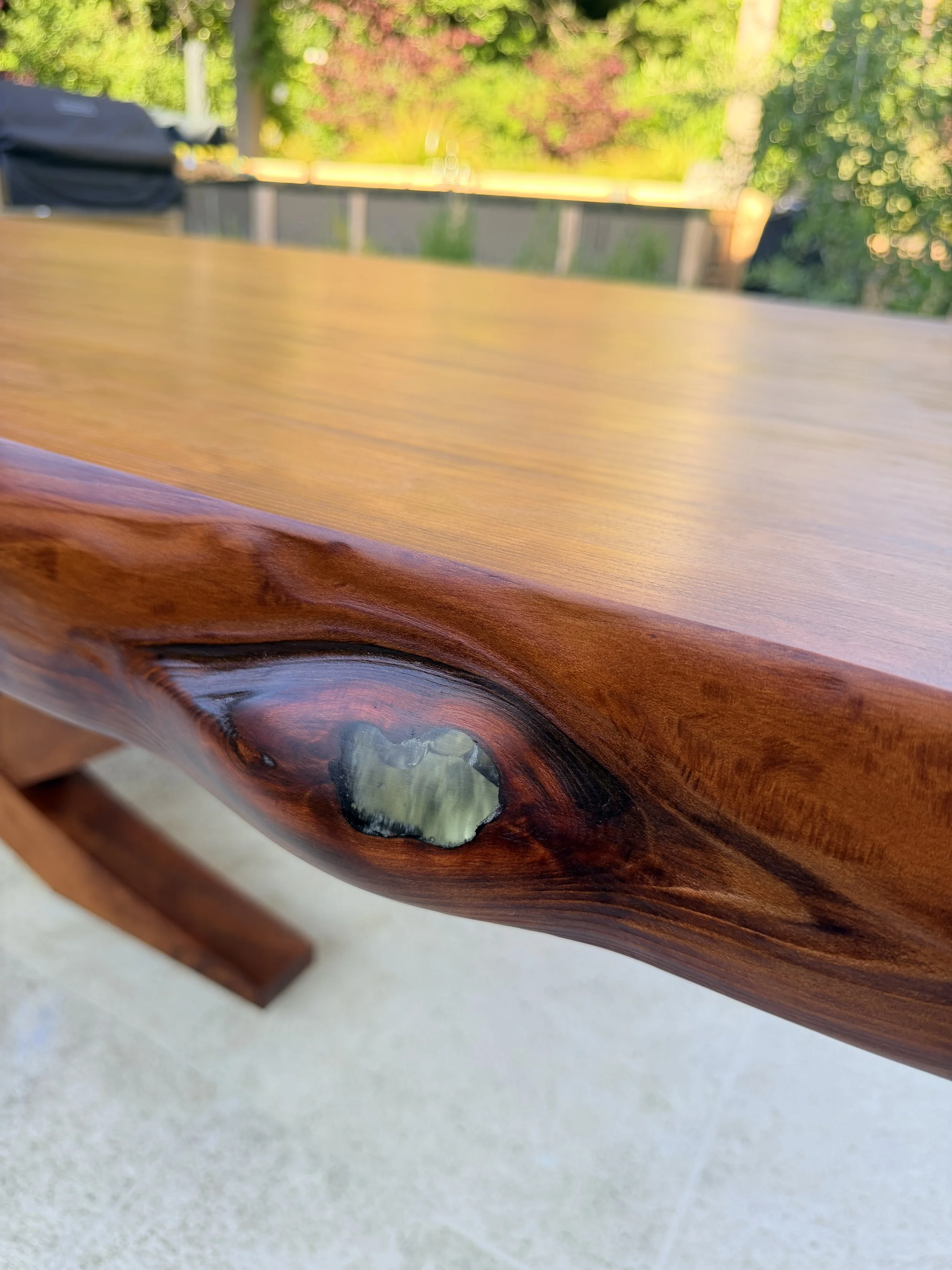 Close-up of a polished wooden table with a live edge and a knot with resin, outdoors with greenery in the background.