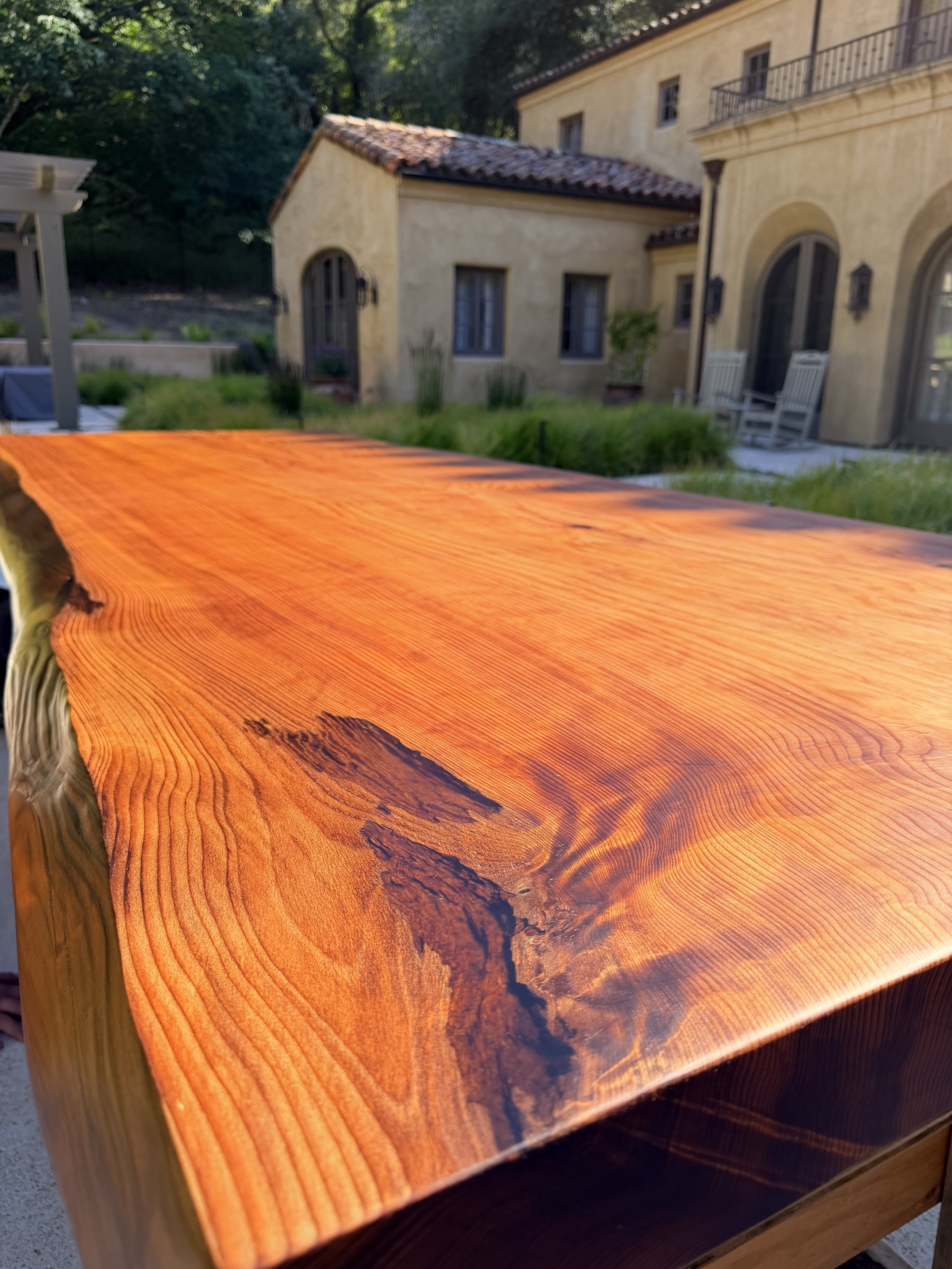 Close-up of a polished wooden table with rich, warm tones and visible wood grain, outdoors in front of a yellow stucco house with arched windows and surrounded by greenery.