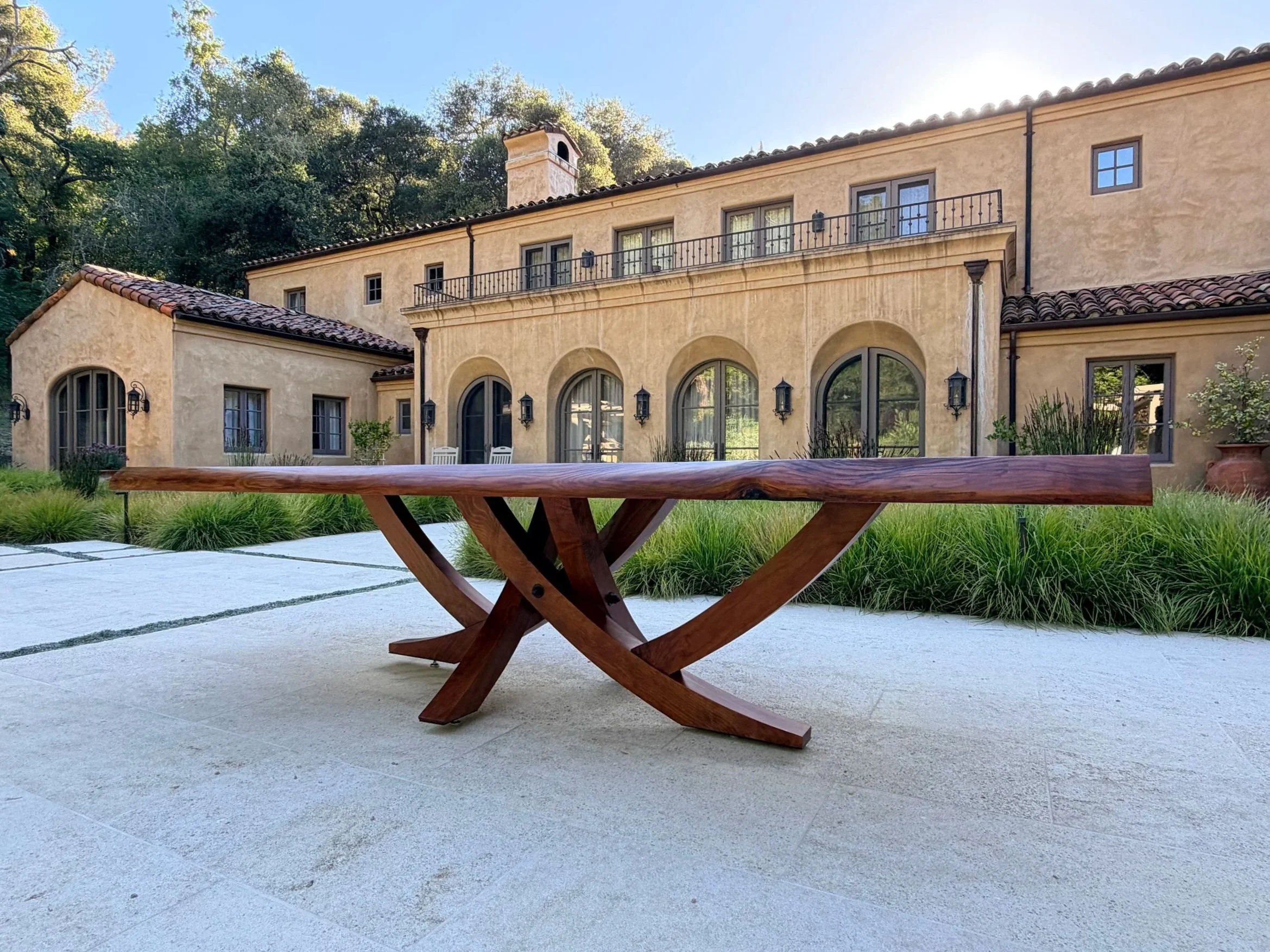 A courtyard with a white stone patio, a wooden outdoor table in the foreground, lush greenery, and a large multi-story house with beige stucco walls, arched windows, and a balcony. The house has dark window frames and a tiled roof, set against a backdrop of trees and a bright blue sky.