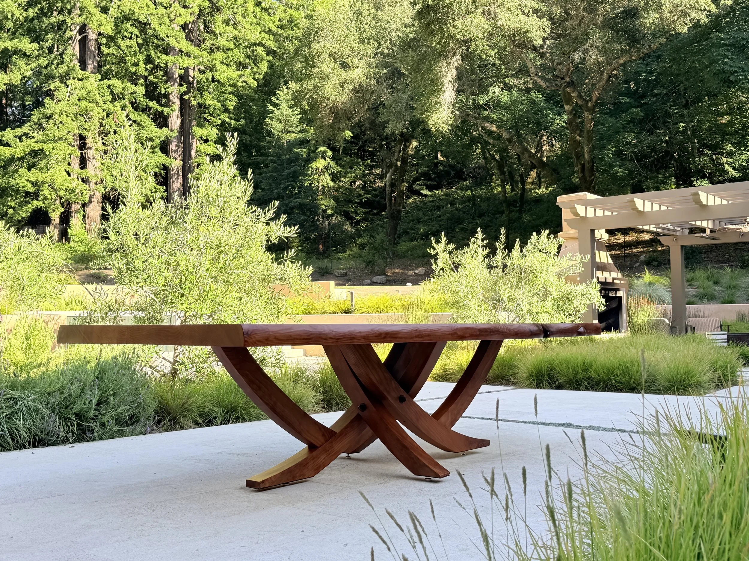 Wooden outdoor table on concrete patio in a lush green garden with trees and plants.
