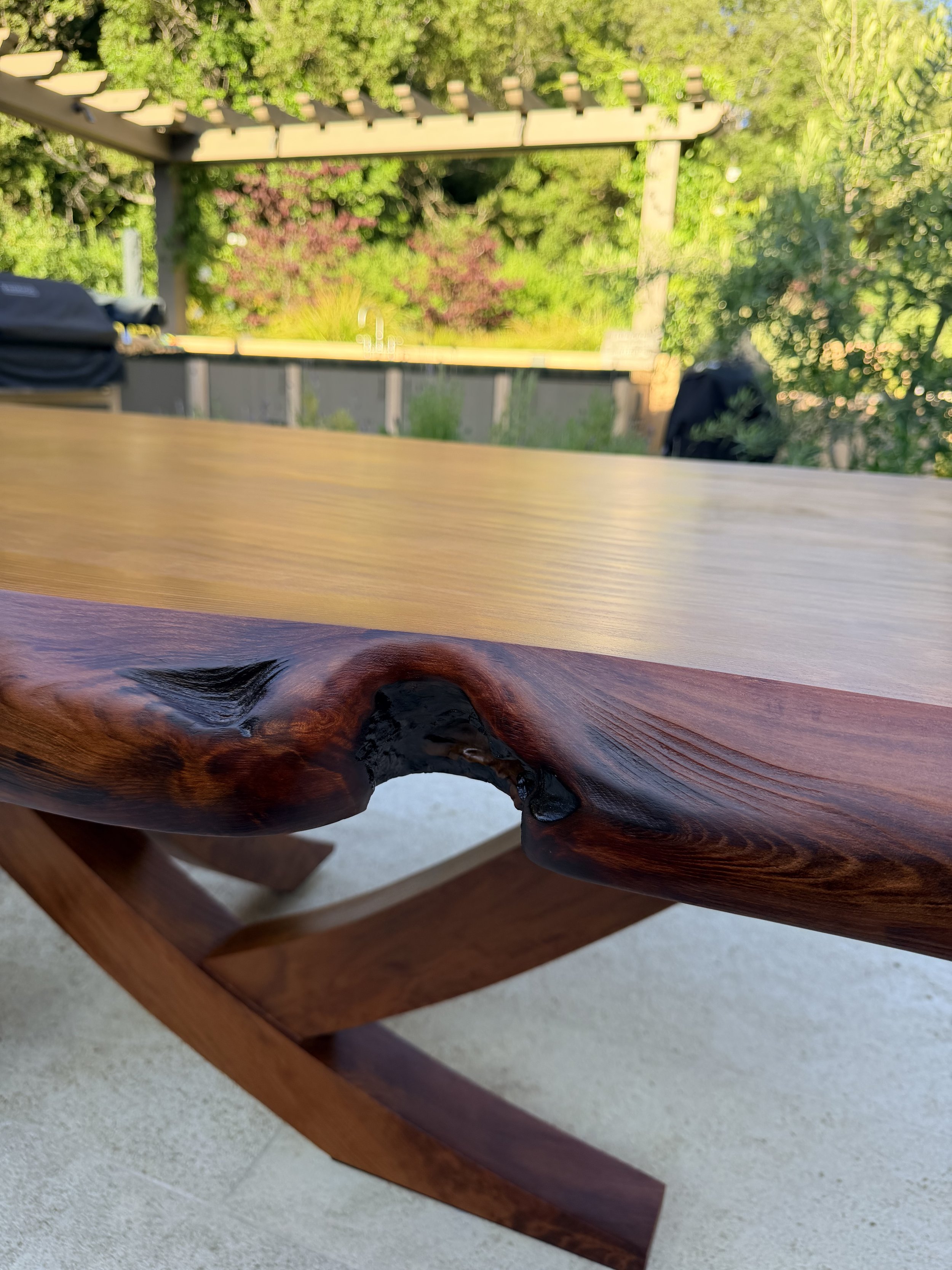 Close-up of a wooden outdoor table with detailed wood grain, part of a wooden chair leg visible, set outside with blurred greenery in the background.