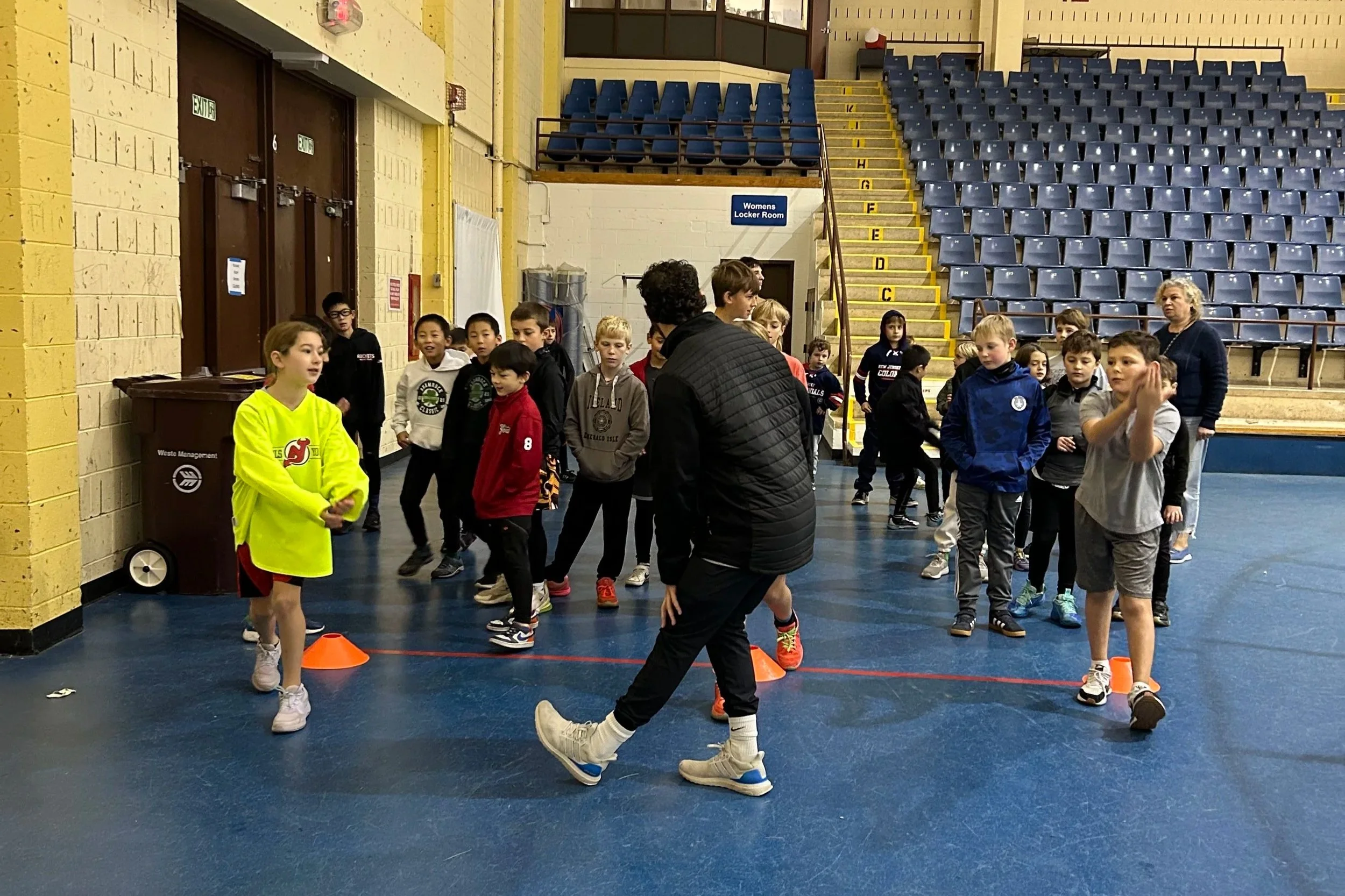 Children in athletic clothing participating in a gym class or sports activity inside a gymnasium with blue flooring, yellow walls, and bleachers in the background.