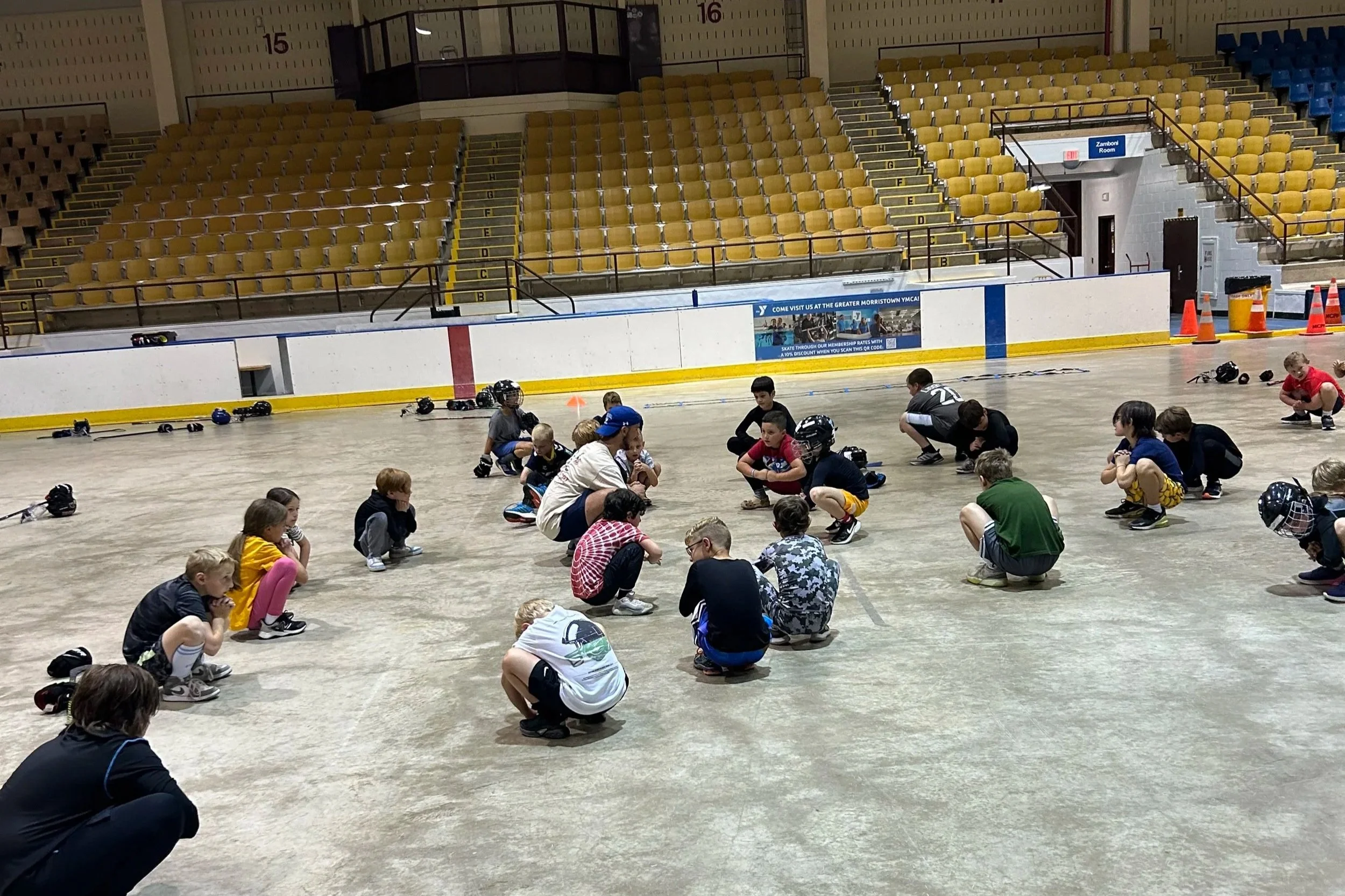 Children on roller skates and helmets during a skate skate lesson at an indoor rink with empty yellow and blue spectator seats in the background.