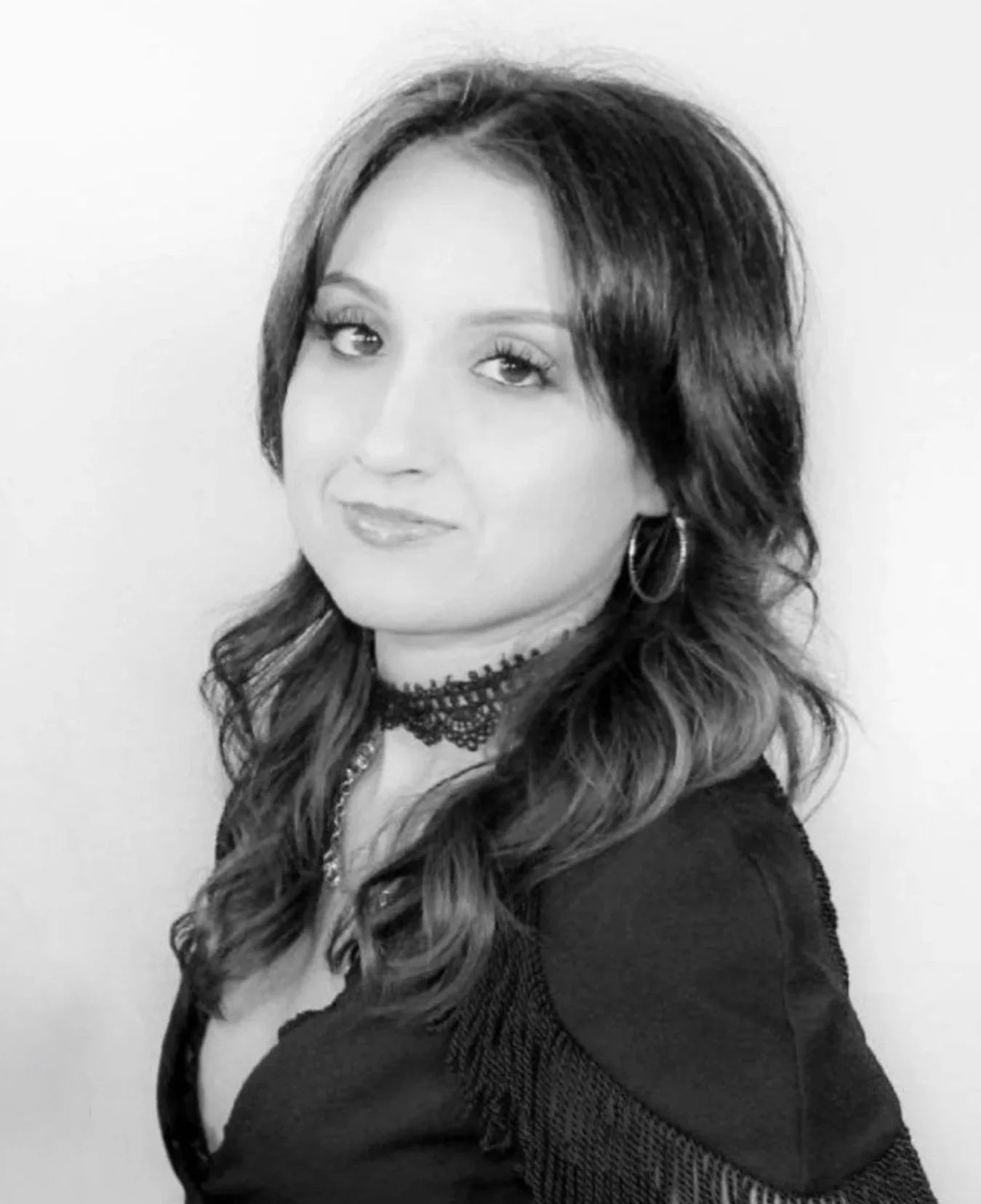 Black and white portrait of a woman with long, wavy hair, wearing earrings, a choker necklace, and a dark top with textured details.