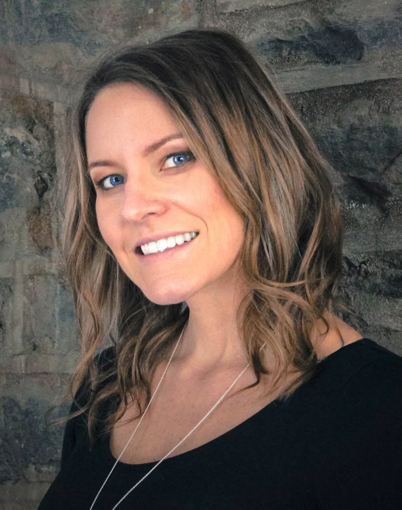 A smiling woman with shoulder-length wavy brown hair and blue eyes wearing a black top and a silver necklace, standing in front of a stone wall.