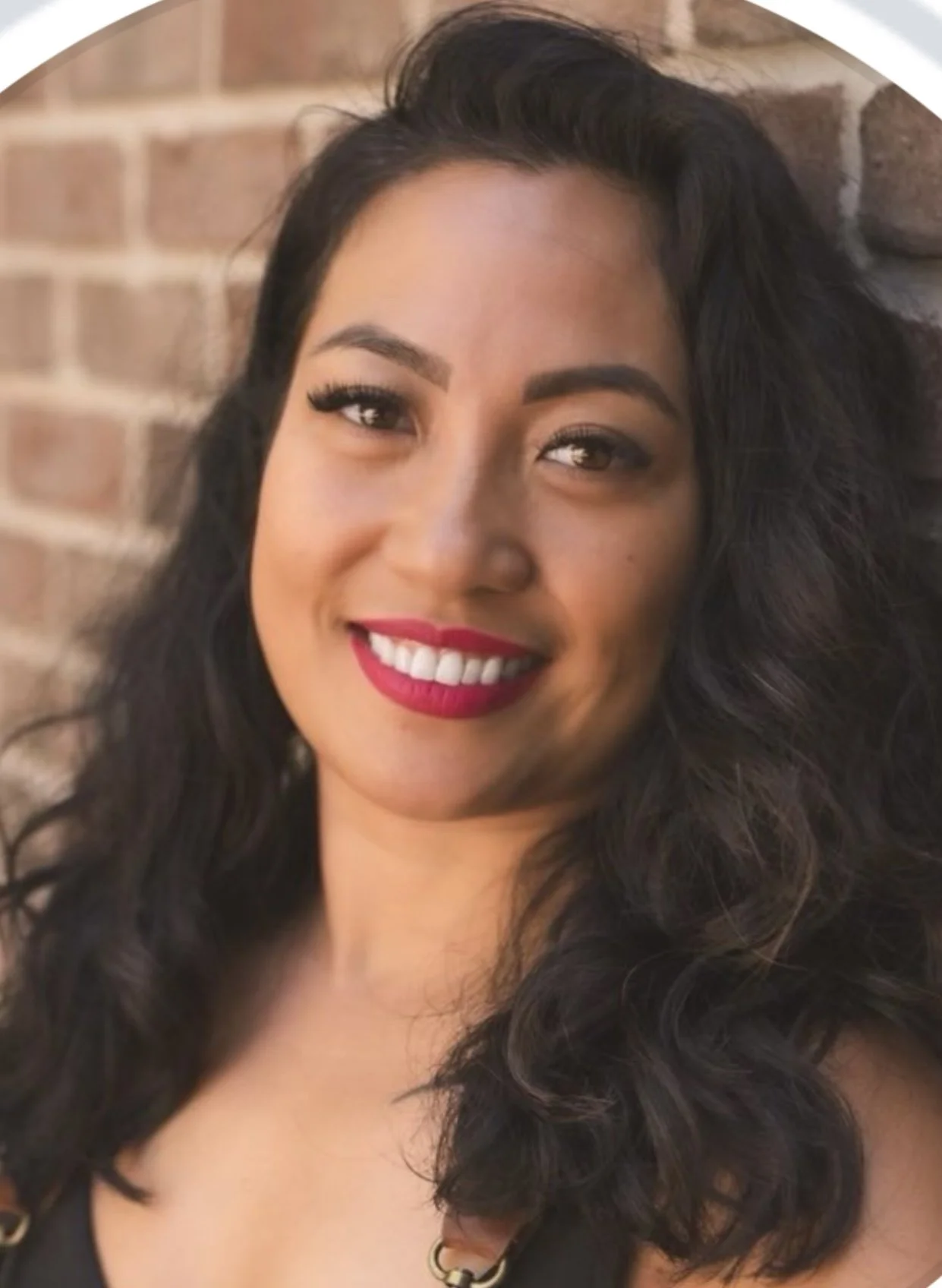 Close-up of a woman with dark, curly hair, wearing red lipstick, smiling in front of a brick wall.