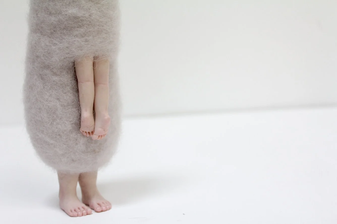 A person in a fuzzy beige sweater standing barefoot on a white surface with a white background, with only their lower legs and feet visible.