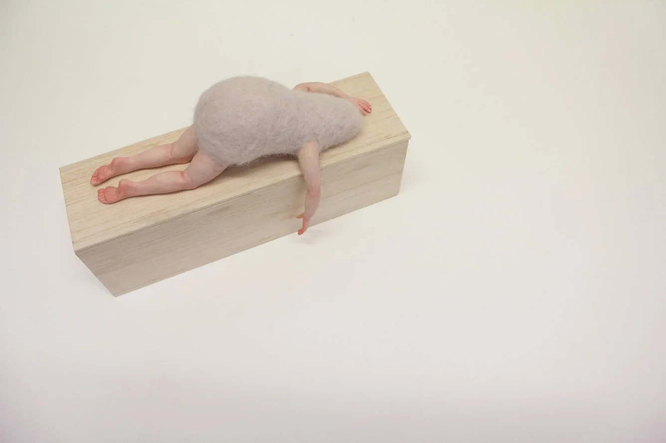 A  felted body and realistic limbs is lying on a wooden block with its belly to the surface, with its hand hanging over the edge.
