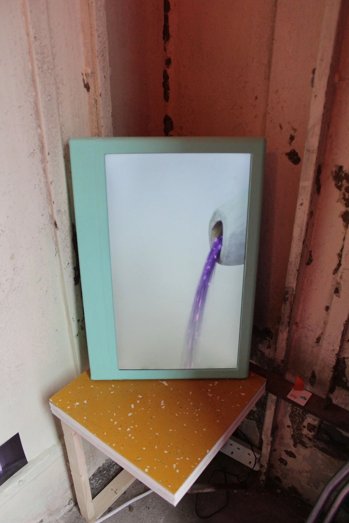 A framed picture of purple water flowing out of a pipe, placed on a small yellow table with a speckled pattern, against a rough wall.