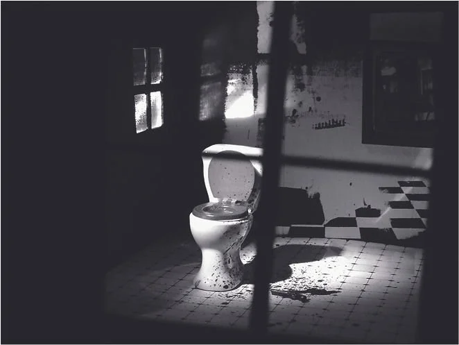 Old, dirty toilet in a dark, abandoned bathroom with broken tiles and a barred window.