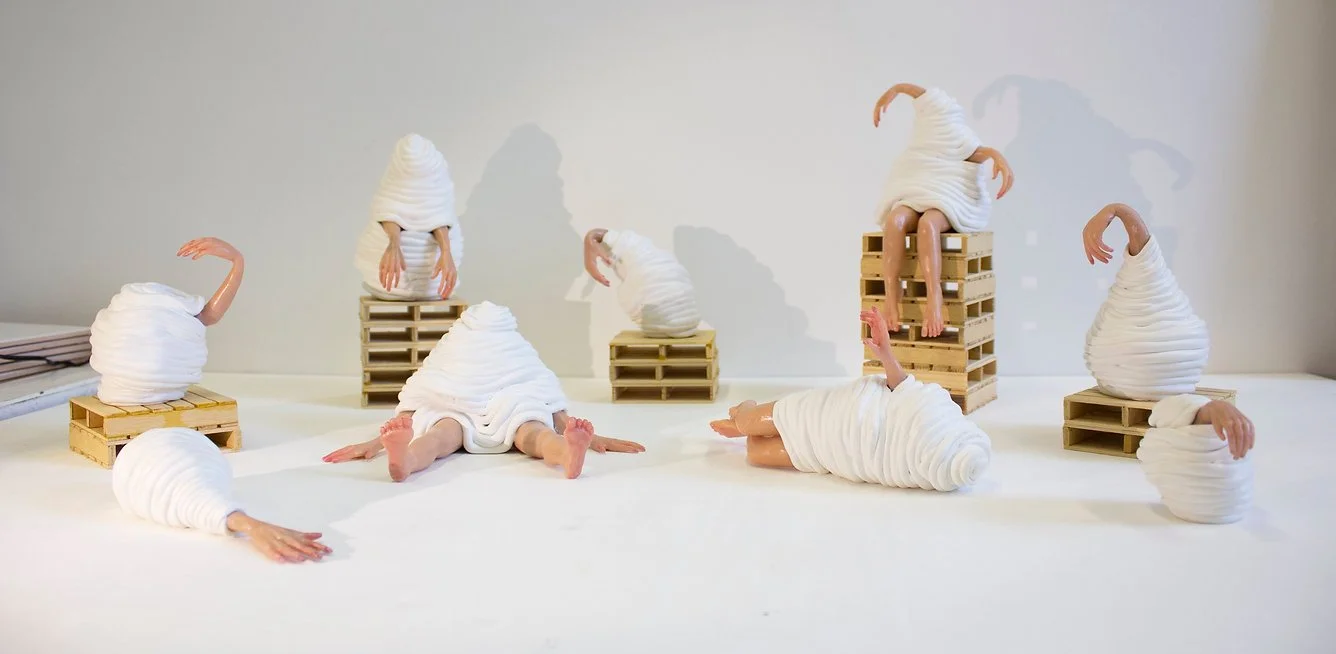 Art installation of whimsical figures wrapped in white fabric, with limbs protruding, arranged on wooden pallets and a white surface, with shadows cast on a plain background.