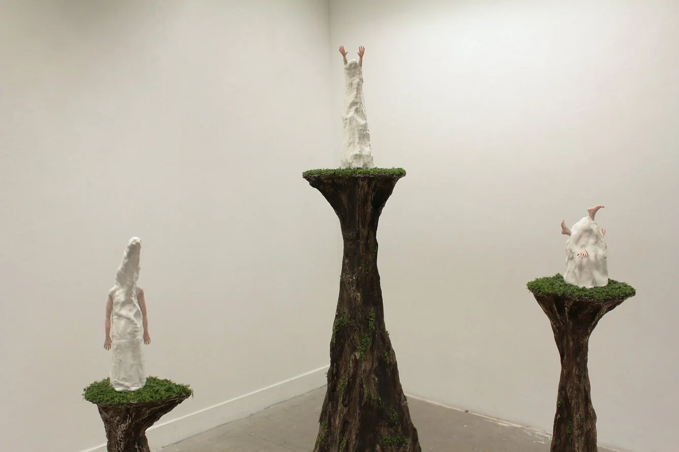 Three artistic sculptures resembling humanoid figures on tall, narrow, tree-stump platforms with green mossy tops, placed in an art gallery with plain white walls.