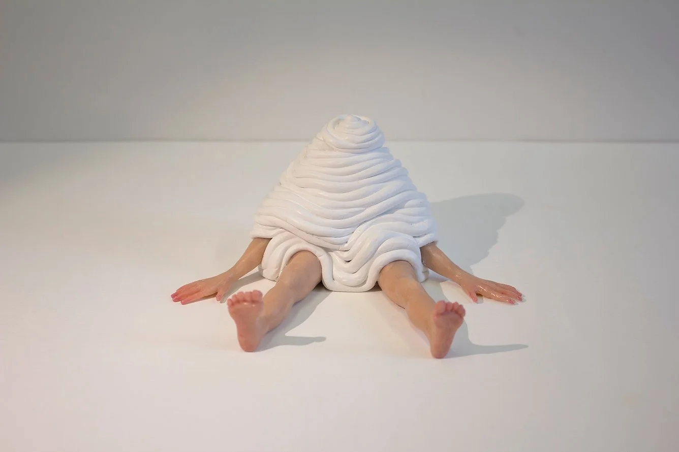 An art piece resembling a human figure with a body covered in white spiral-shaped material, lying face down with arms and legs extended on a plain white surface.