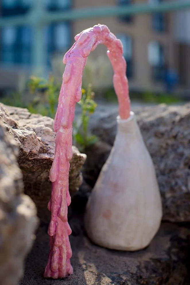 Pink waterfall with wax-like texture flowing from a white ceramic vase onto rocks, set outdoors with a blurred urban background.