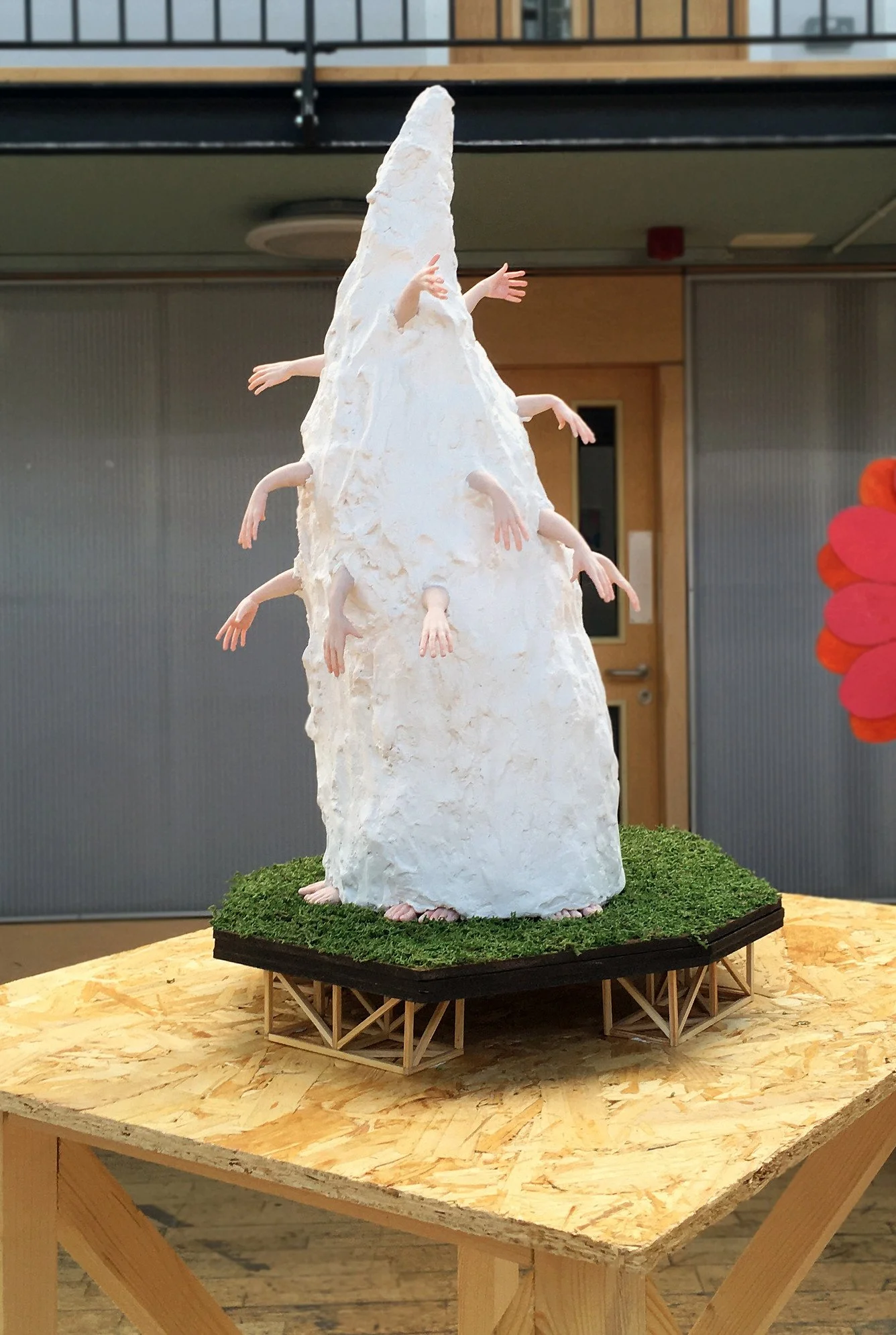 An art sculpture of a person covered in a white substance resembling snow or ice, with multiple human arms protruding from the main body, displayed on a grassy base on a wooden table.