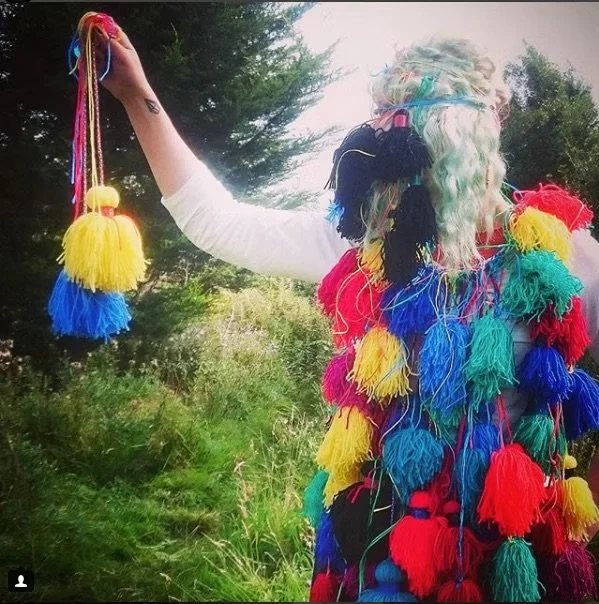Person wearing a multicolored, tassel-covered costume with a mask of a person's face, standing outdoors in a green landscape with trees and shrubs.