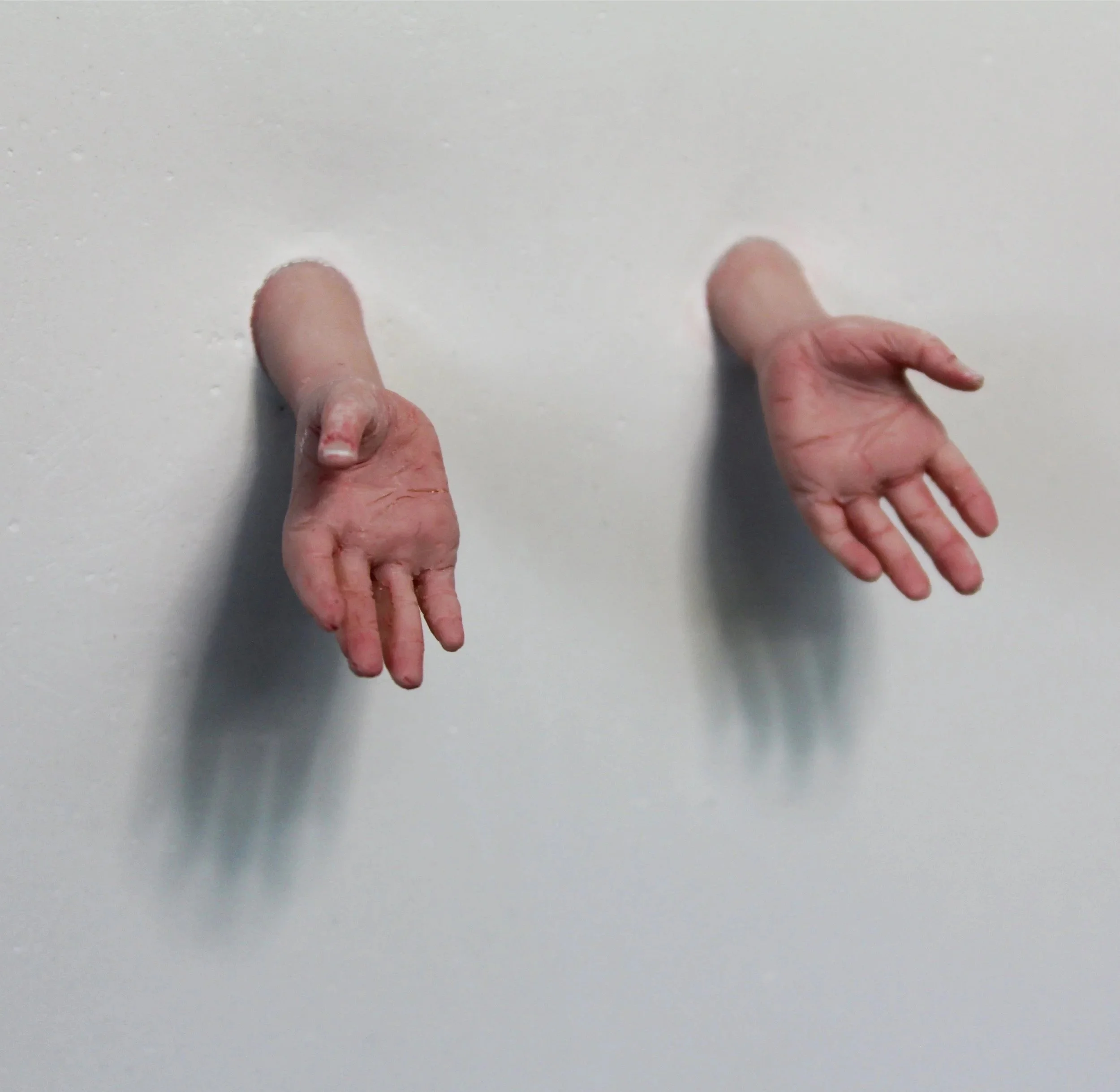 Two human arms protruding from a plain white wall, with hands reaching outward.