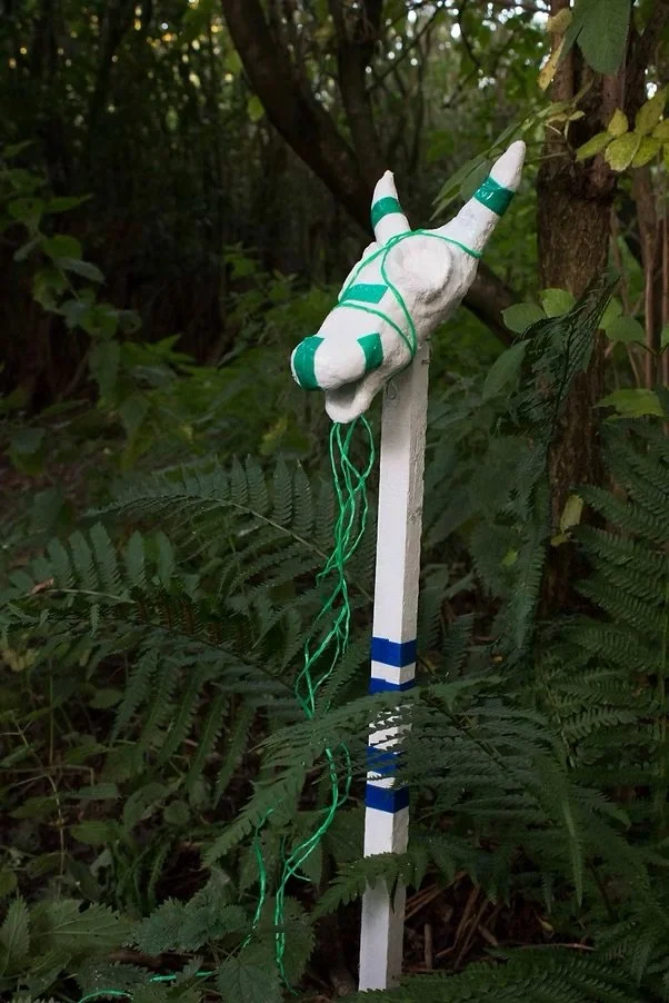 A sculpture of white animal like head with teal accents, hanging from a white and blue striped wooden stake amid greenery.