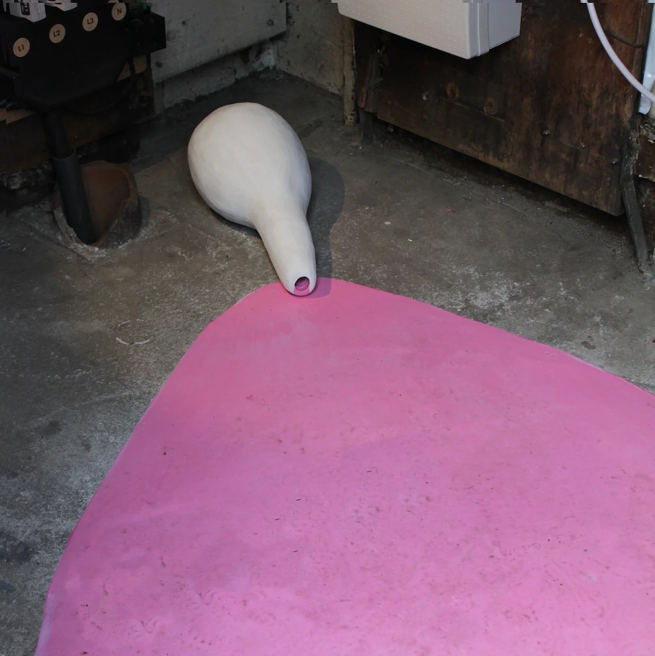 A large, white, handmade sculpture  lying on a cracked concrete floor. The sculpture  is located next to a pink-painted area on the ground, possibly part of an art installation. 