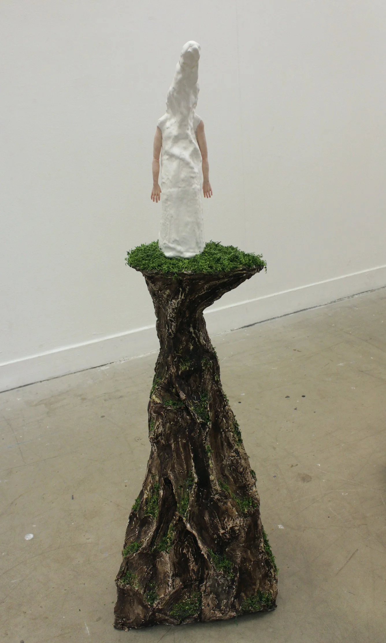 An art installation with a wooden stump base, a small patch of green moss, and a figure with a white form covering the upper body, creating an abstract, surreal sculpture.