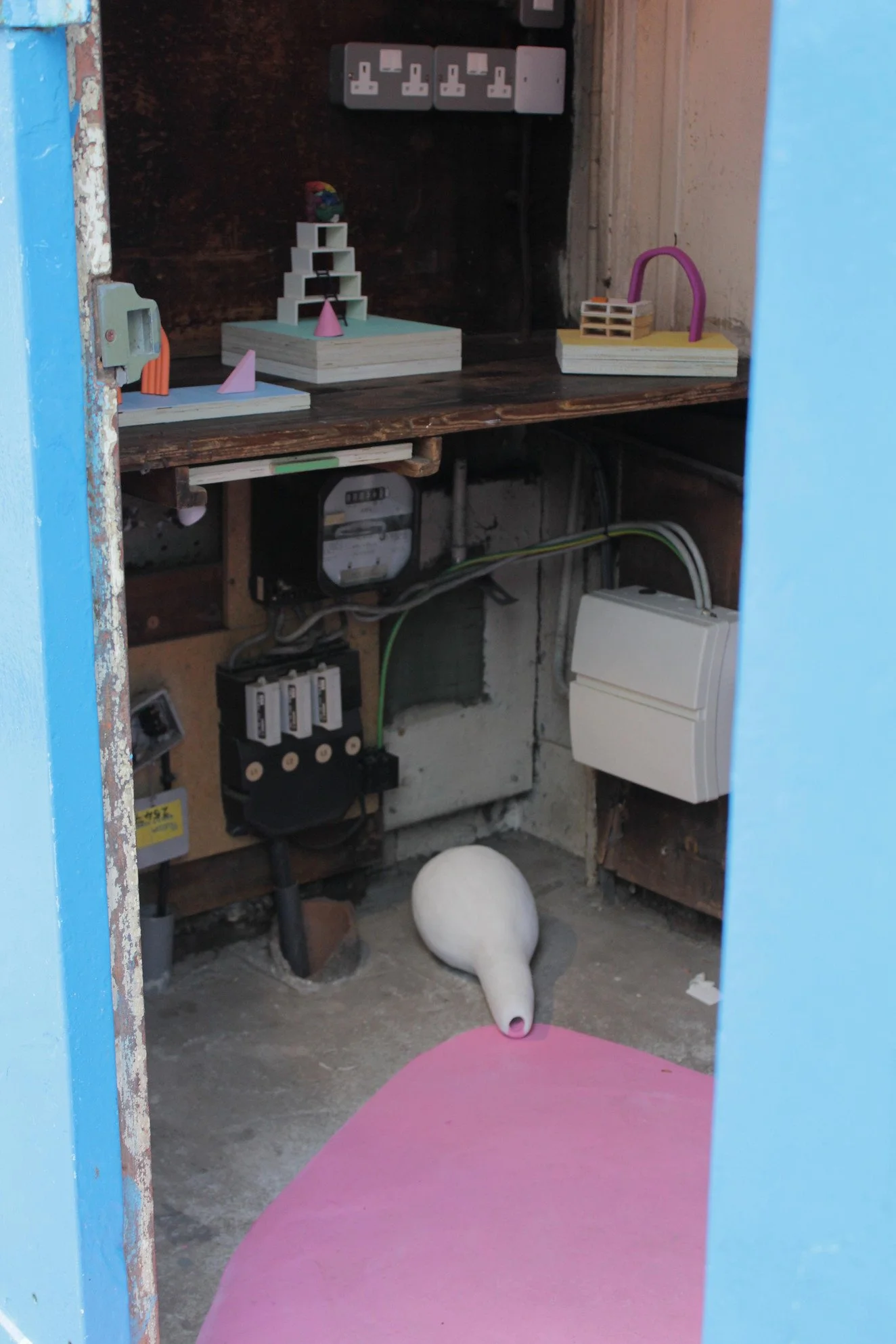 View inside a utility or storage room with a pink mat on the floor, a white ceramic watering can, and wooden shelves holding small geometric sculptures and toys. Electrical outlets and wiring are visible on the wall.