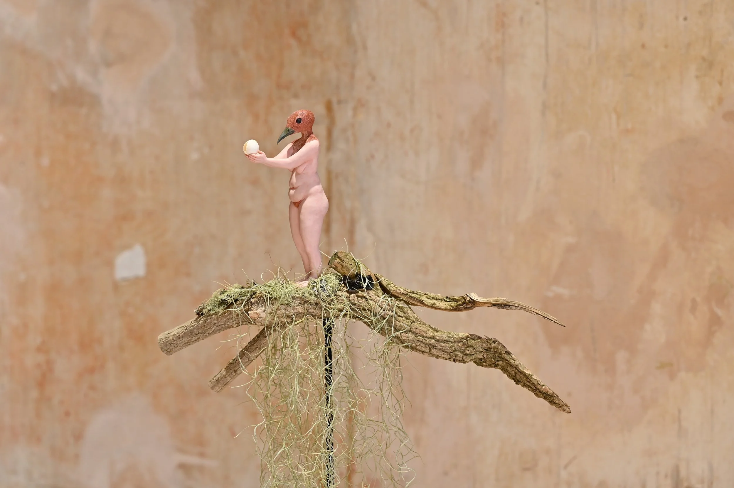 A surreal sculpture of a human figure with a bird's head, holding a small egg, standing on a tree branch with moss and vines, against a textured beige background.