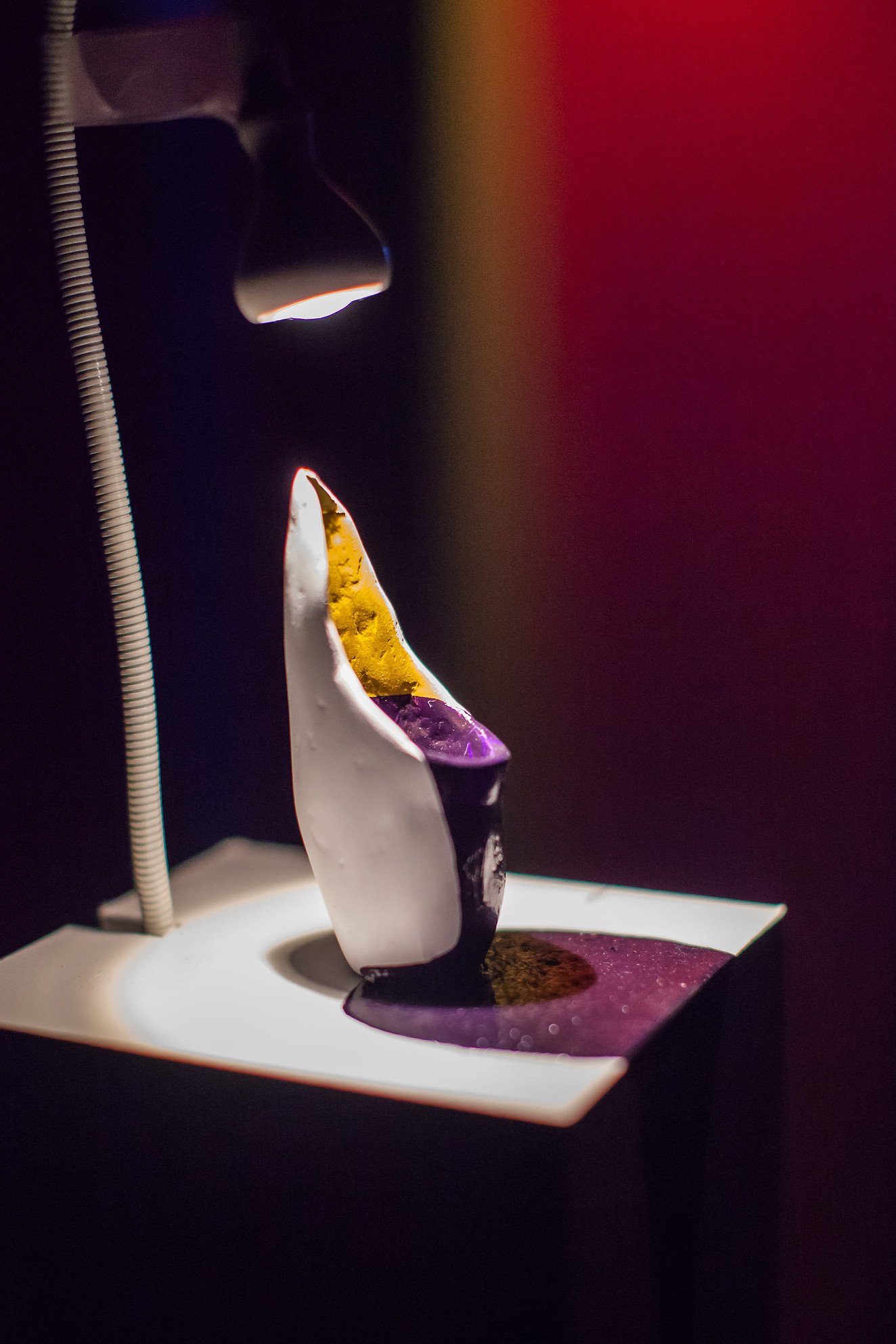 A modern abstract sculpture with white, yellow, and purple sections, lit by a focused light, displayed on a pedestal in a dark gallery setting.